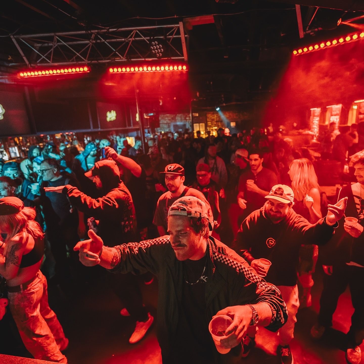 Crowded indoor party with red and blue lighting; people are dancing, holding drinks, and enjoying the lively atmosphere under bright stage lights.