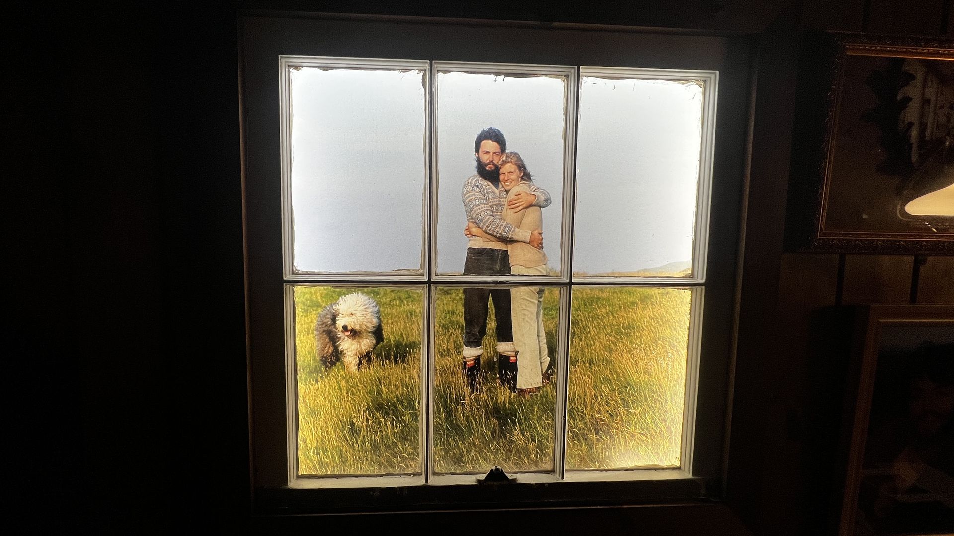 Window with eight glass panes showing a photo: a bearded man and a woman hug in the center, wearing sweaters; a fluffy dog stands in a sunlit grassy field at the bottom; blue sky behind.