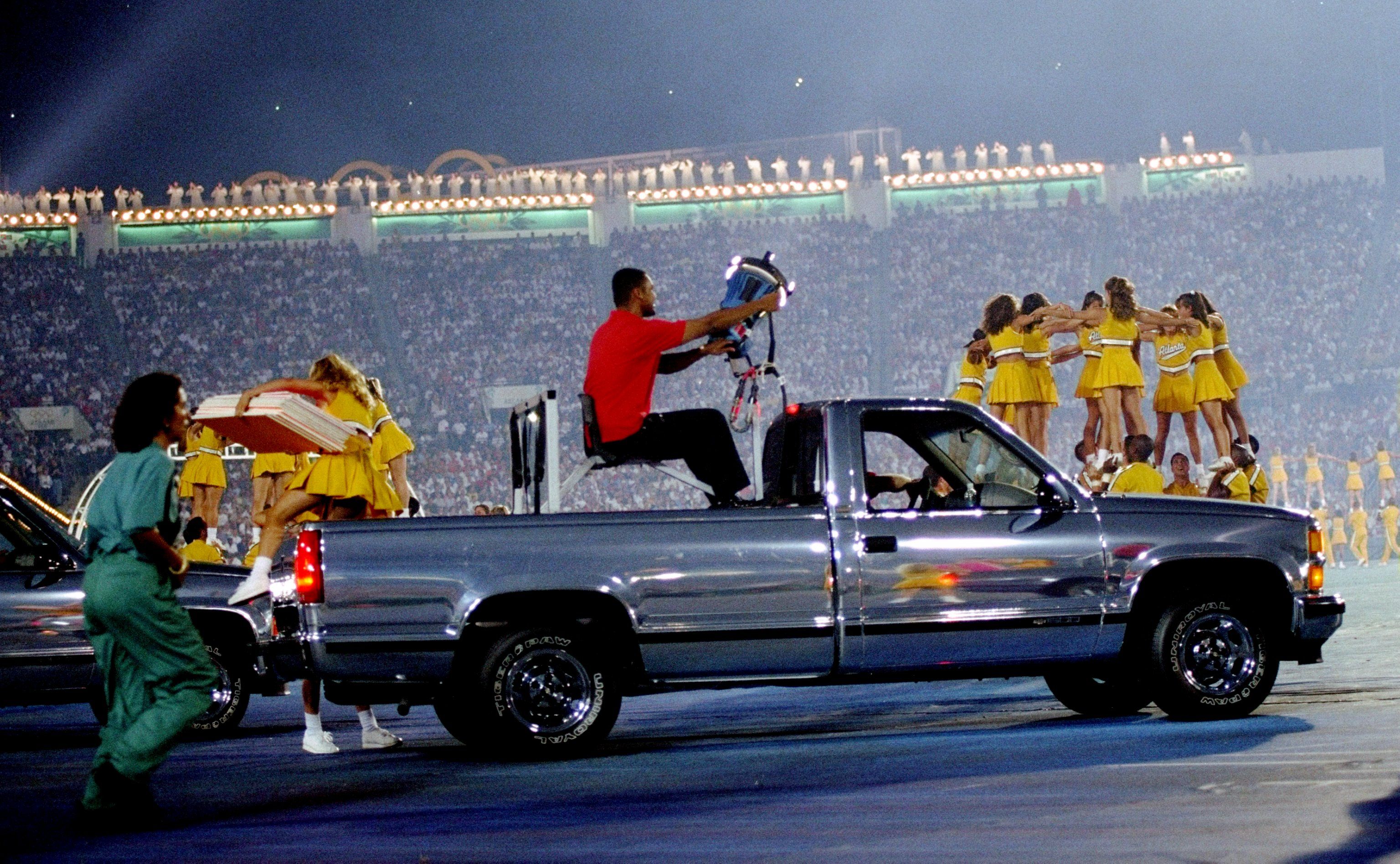 A chrome pick-up truck with a large light drives past a group of cheerleaders performing a pyramid in a packed stadium