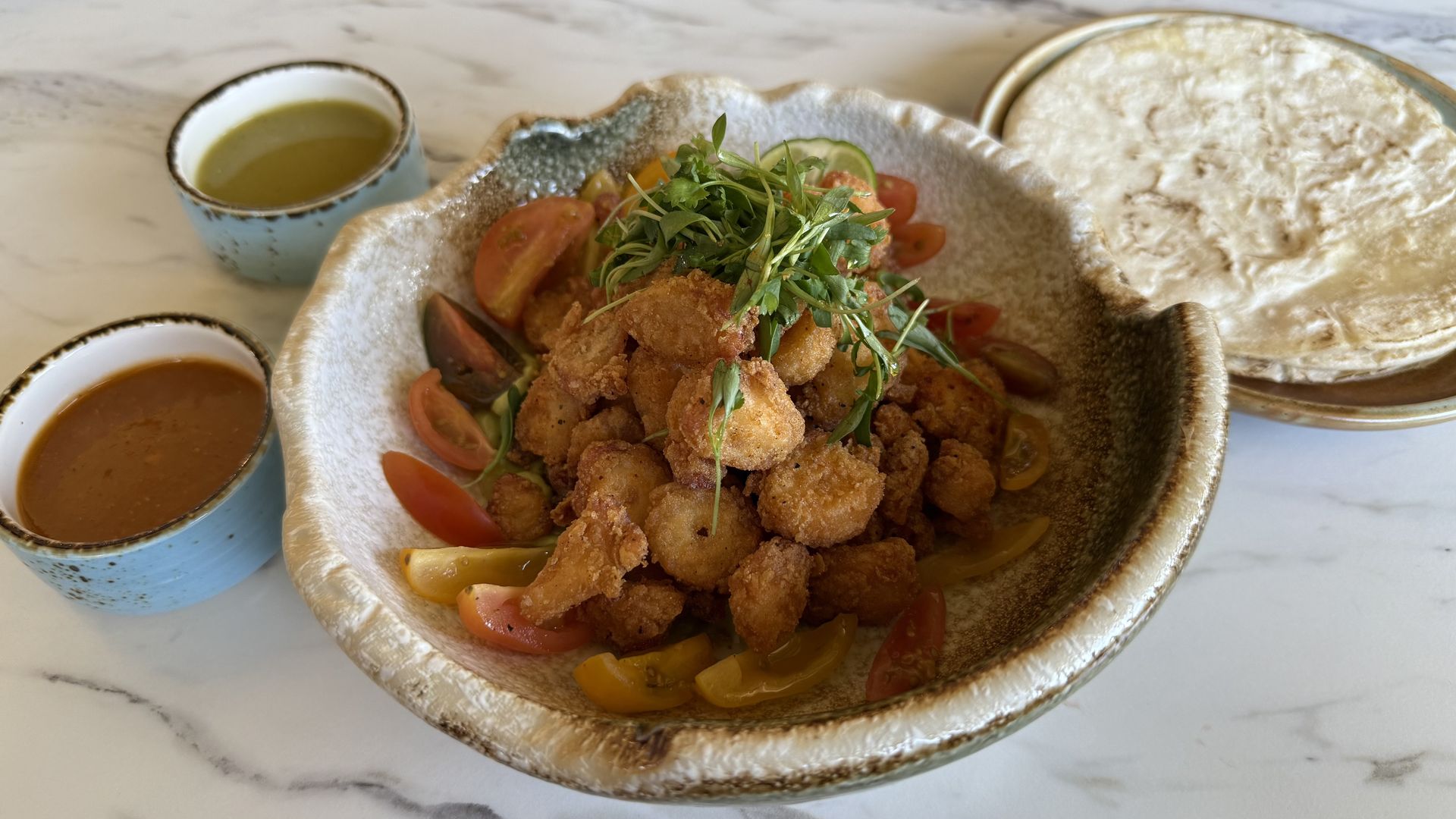 A ceramic bowl filled with fried and breaded nuggets of octopus with greens on top. Next to the bowl are two cups of red and green sauce, and a plate of small tortillas.