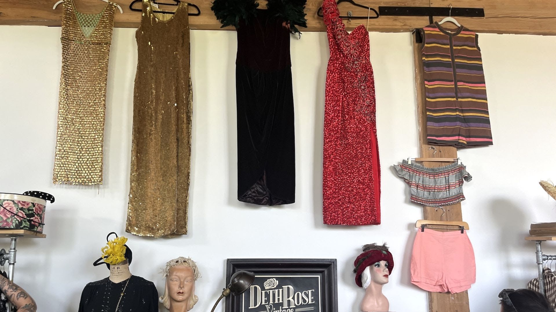 Five dresses and outfits hanging on a wooden beam: two gold sequin dresses, one black dress, one red sequin dress, and a striped sleeveless top with matching shorts below with a sign that reads DethRose
