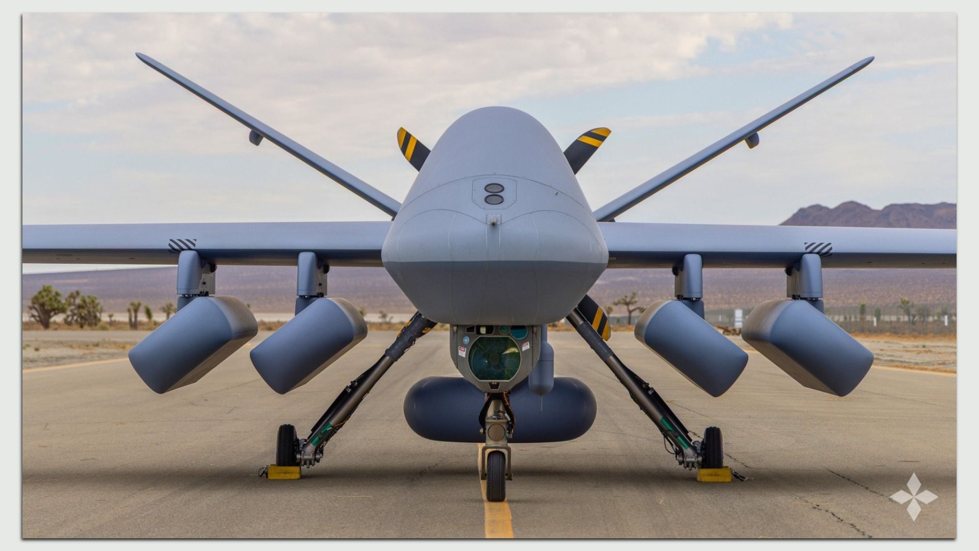 Front view of a gray unmanned aerial vehicle on a desert runway, with wide wings, multiple underwing pods, a bulbous sensor nose, visible landing gear, and distant mountains under a cloudy sky.