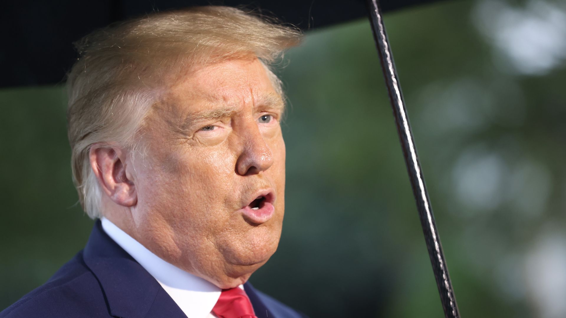 Trump, wearing a suit and tie, speaks to reporters while holding an umbrella