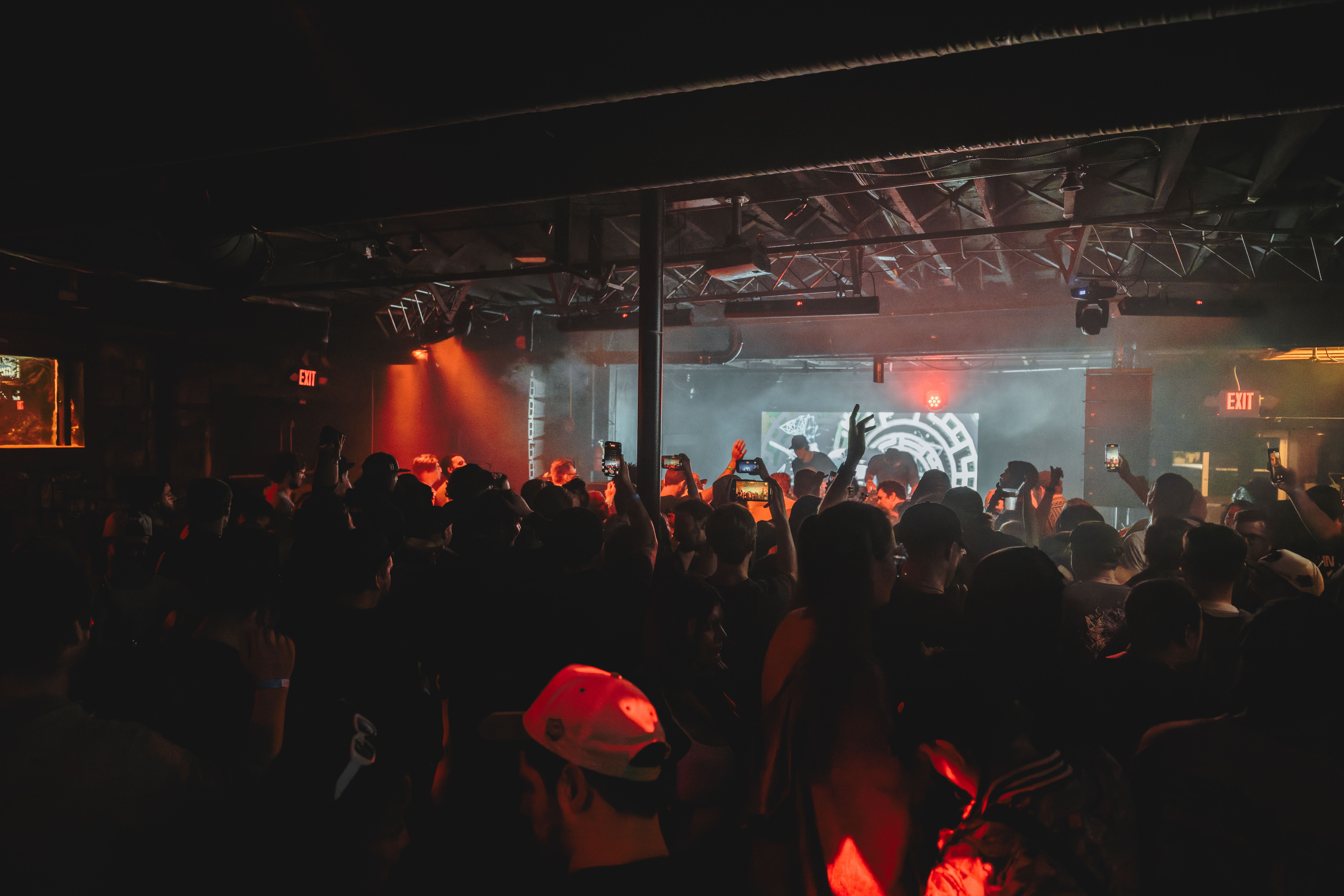 Crowd of people at a dark indoor concert or club with red and blue lighting, many holding up phones to record. Exit signs glow in the background above the stage.