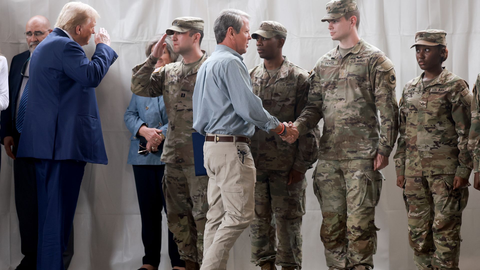 Men and women dressed in military fatigues shake hands with Gov. Brian Kemp and salute U.S. President Donald Trump.