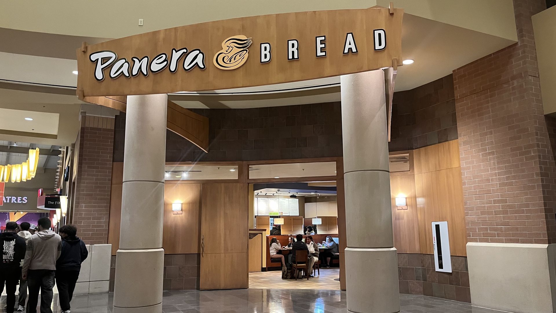 Entrance of Panera Bread inside a mall with wooden panels and pillars; a group of people dining inside; people walking by on the left.