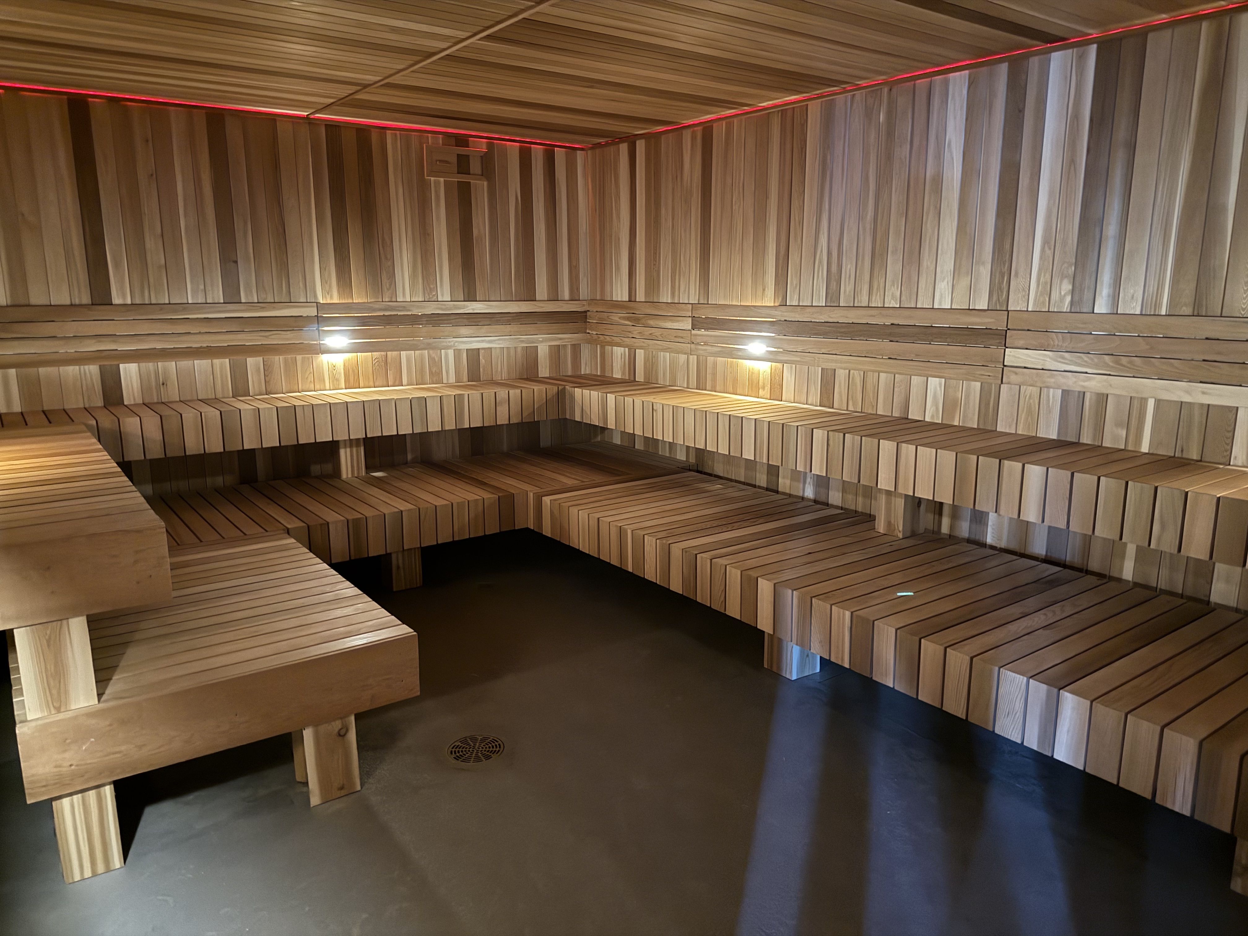 Interior of a wooden sauna with tiered benches along the walls, warm lighting, and a red LED strip near the ceiling, featuring a clean, minimalist design.