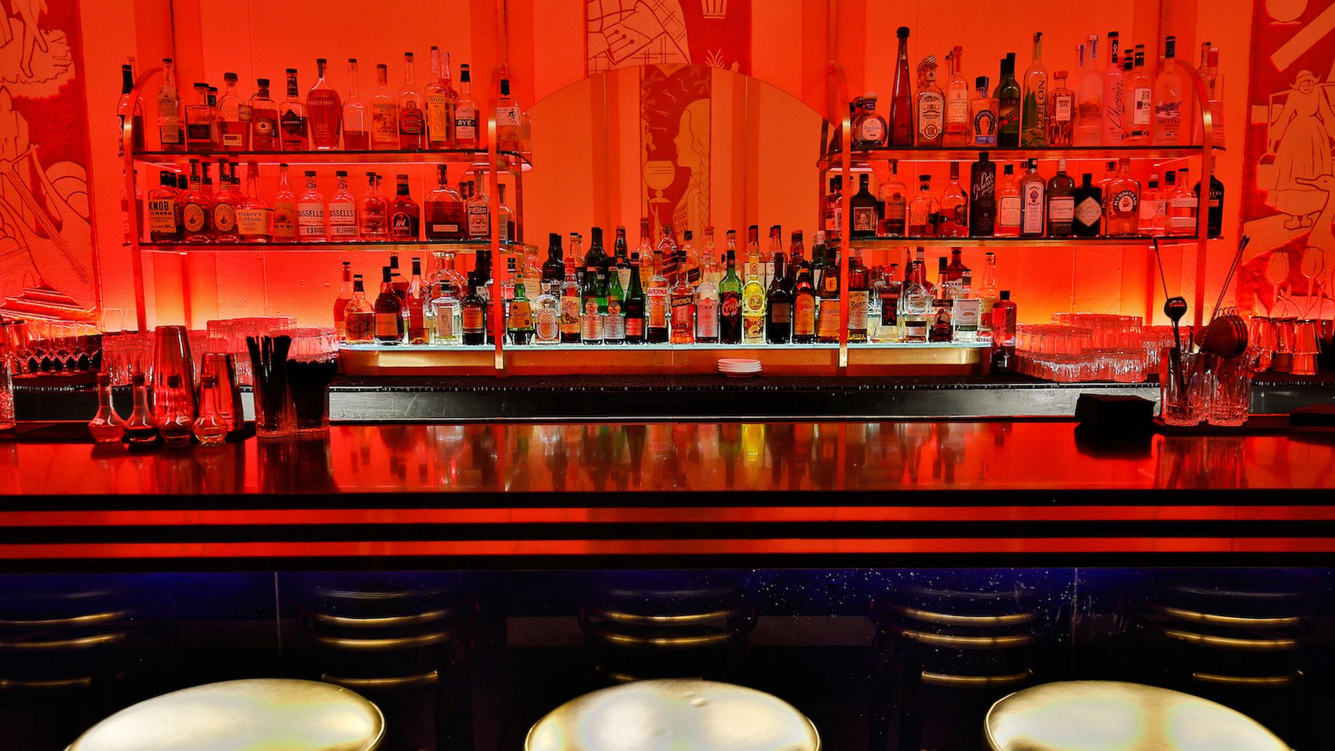 Bar with red-orange lighting, three gold cushioned stools, shelves stocked with various liquor bottles, glassware, and mirrored backdrop featuring abstract artwork.