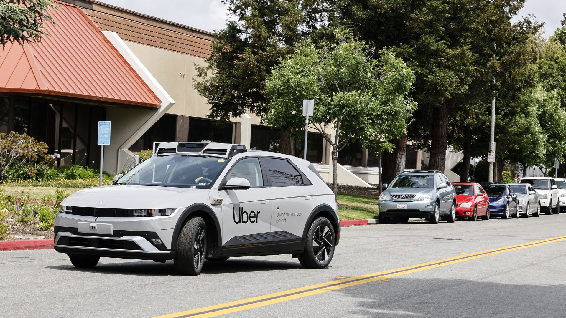 White Hyundai Ioniq 5 SUV with Uber logo on the side, and various sensors on the roof and sides of the vehicle, drives along a city street in front of a beige building with an orange slanted roof; a line of parked cars and lush green trees line the curb.