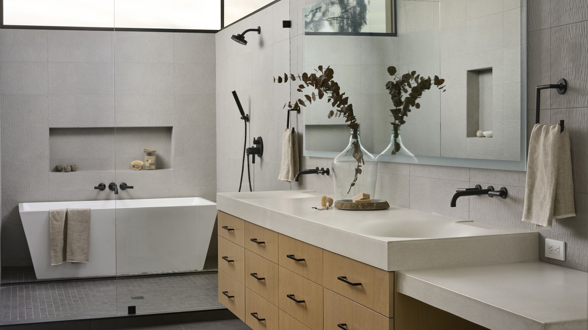 Modern bathroom with a white freestanding tub, black shower fixtures and light wood vanity with white countertop. A partial glass wall separates the tub and shower from the rest of the space.