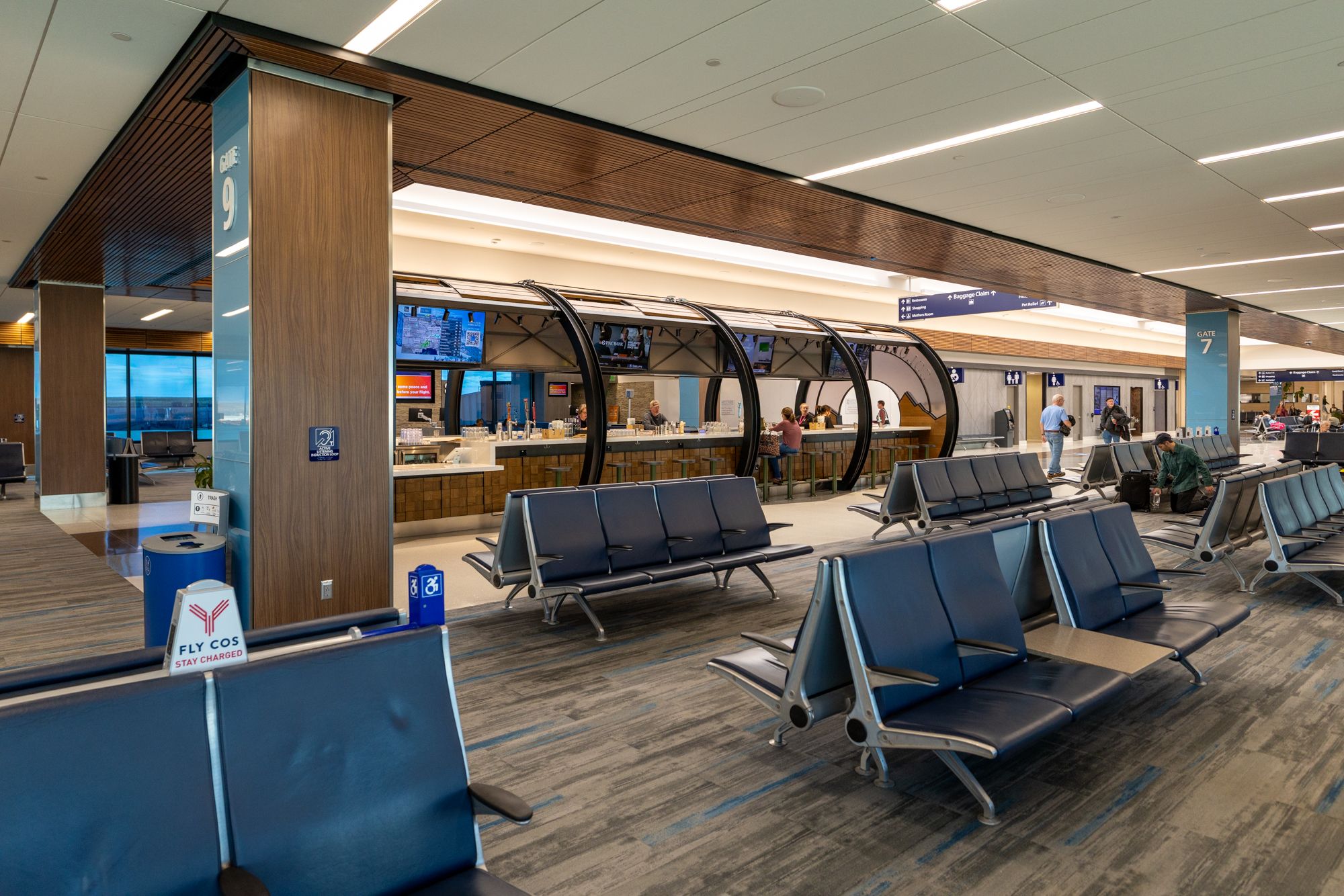 Interior of an airport terminal with blue seating, wood-paneled columns, and curved black arches over service counters. Signage shows Gate 7 and Gate 9; passengers wait and staff assist.