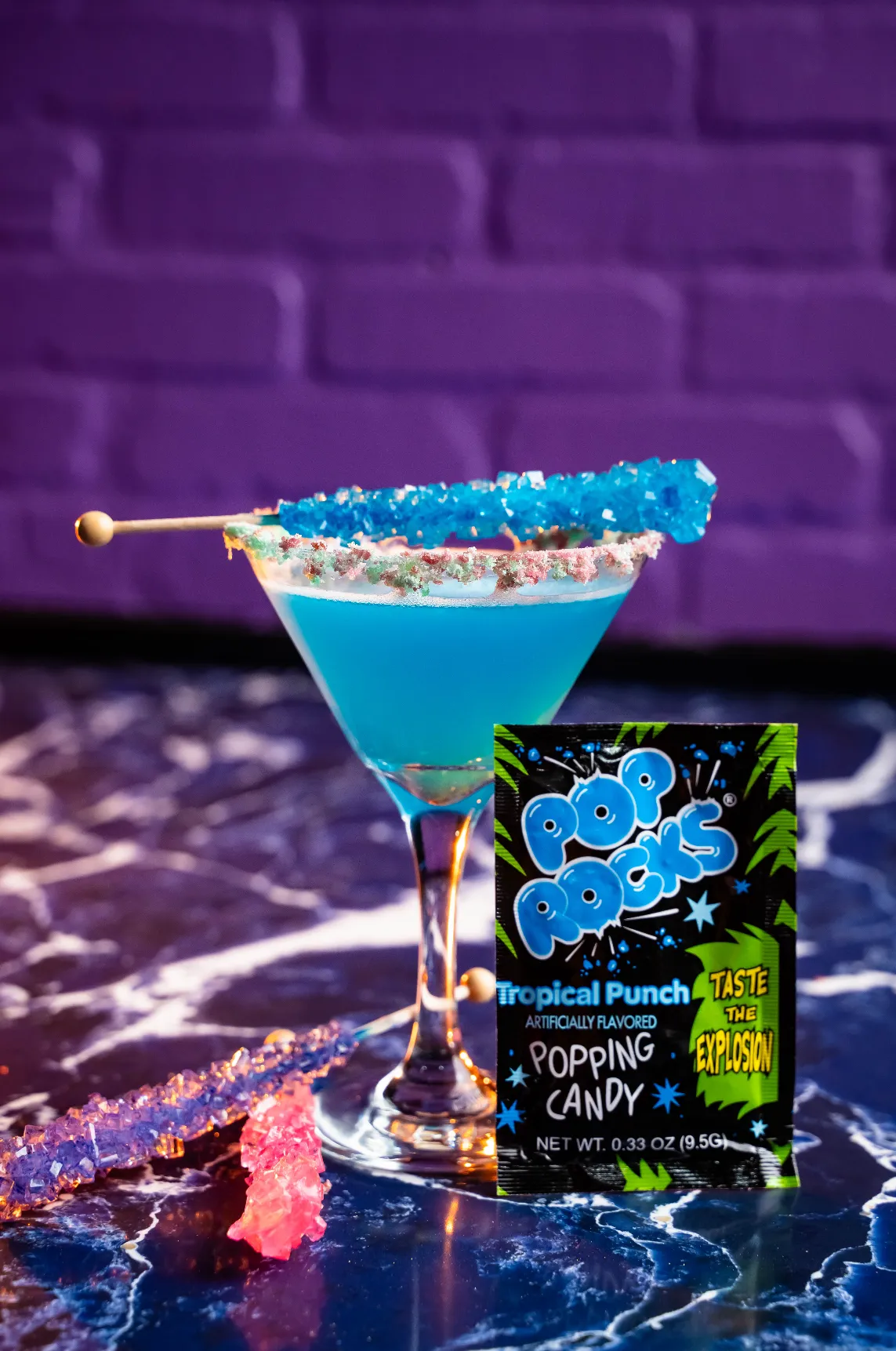 Blue tropical punch cocktail in a martini glass with a colorful sugar rim and a blue rock candy stick on top, next to a packet of "Pop Rocks" popping candy and additional rock candy sticks.