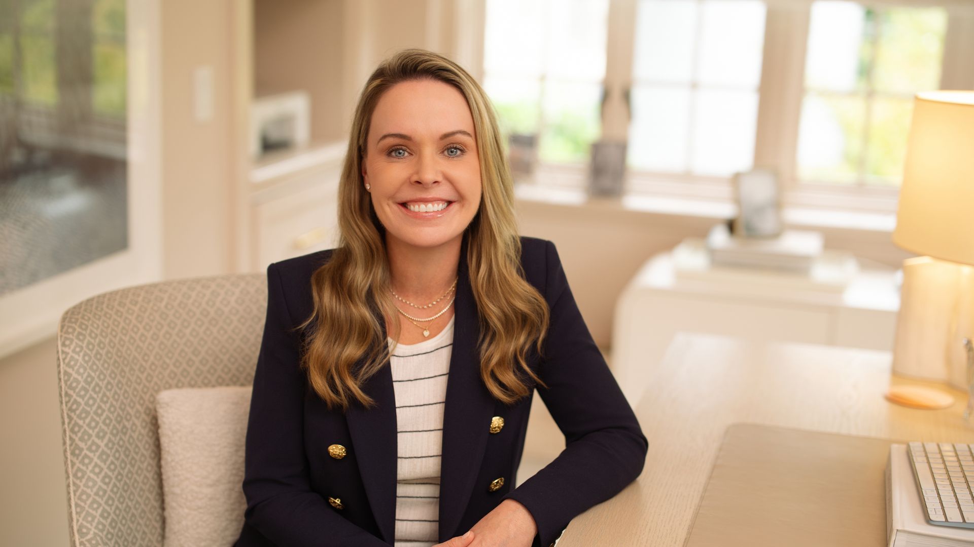 Jill Hazelbaker, a smiling blonde woman in a navy blazer and striped top sits at a desk in a bright office, with a keyboard and a lamp nearby and large windows behind.