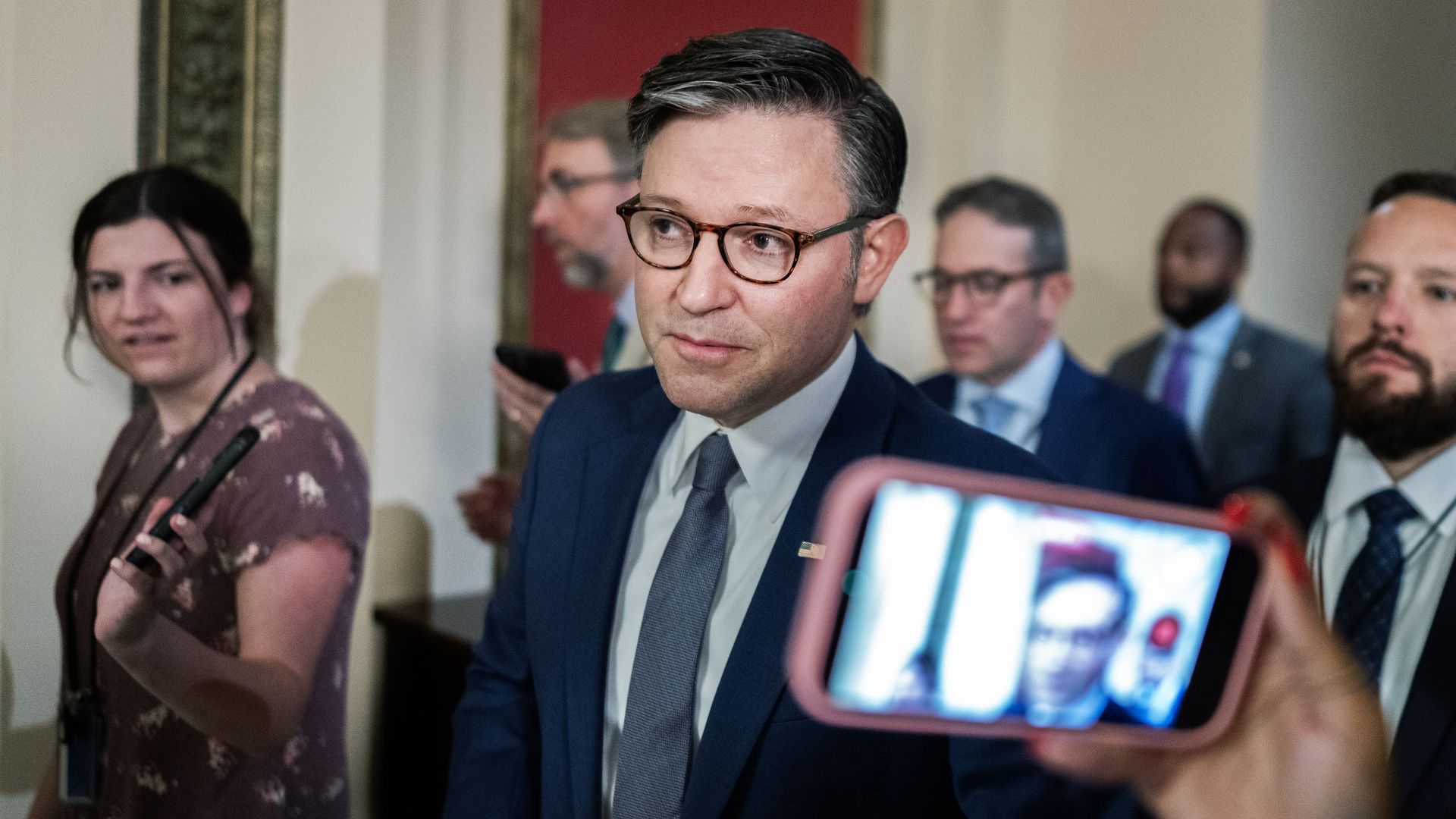 A man in a navy suit and gray tie wearing round tortoiseshell glasses, in a crowd of reporters. A woman in a floral dress holds a microphone, while others watch and record with phones.
