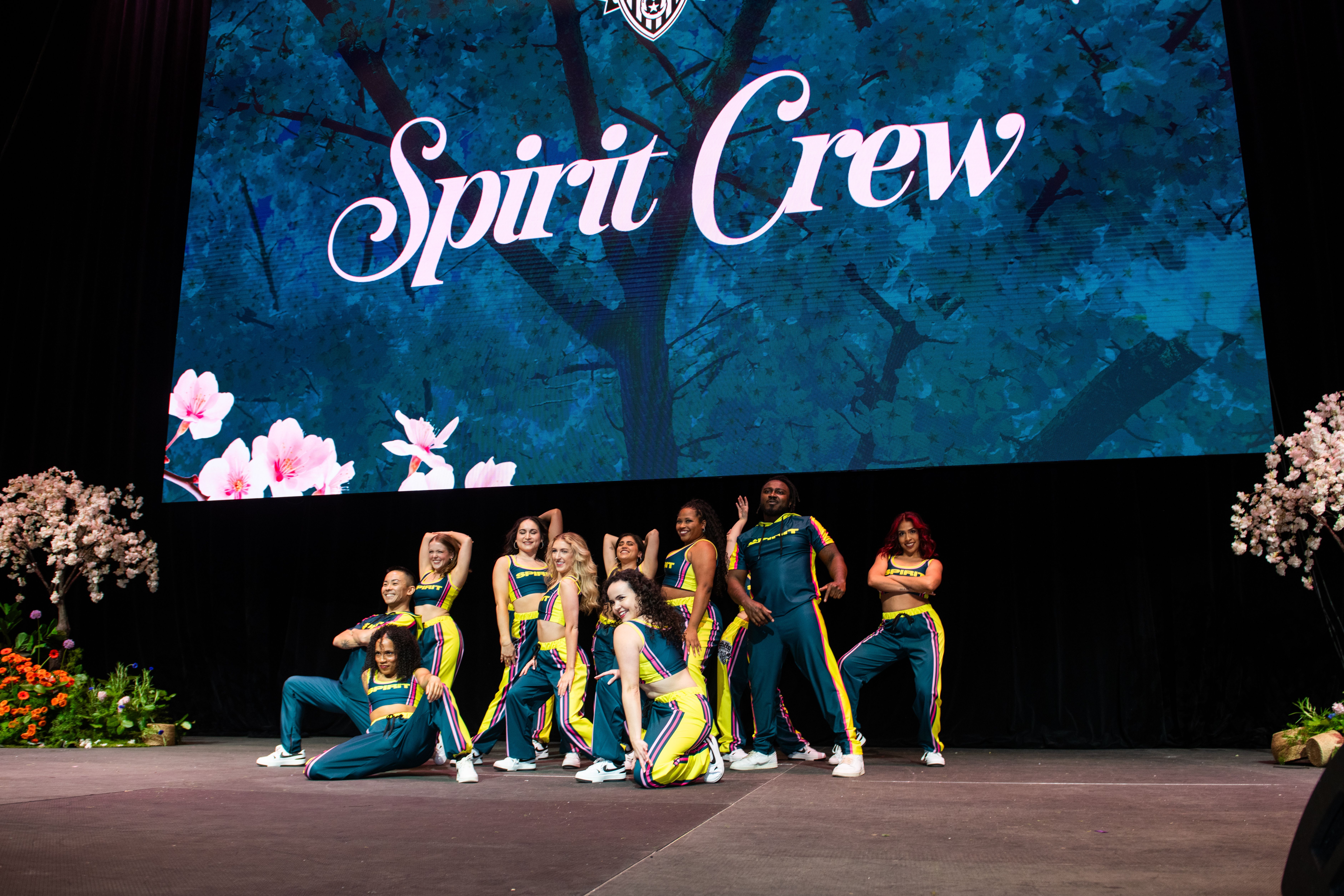 A dance crew performs a routine on stage in teal and yellow outfits with purple stripes, in front of a large backdrop that reads "Spirit Crew" in cursive; floral decorations on the sides.