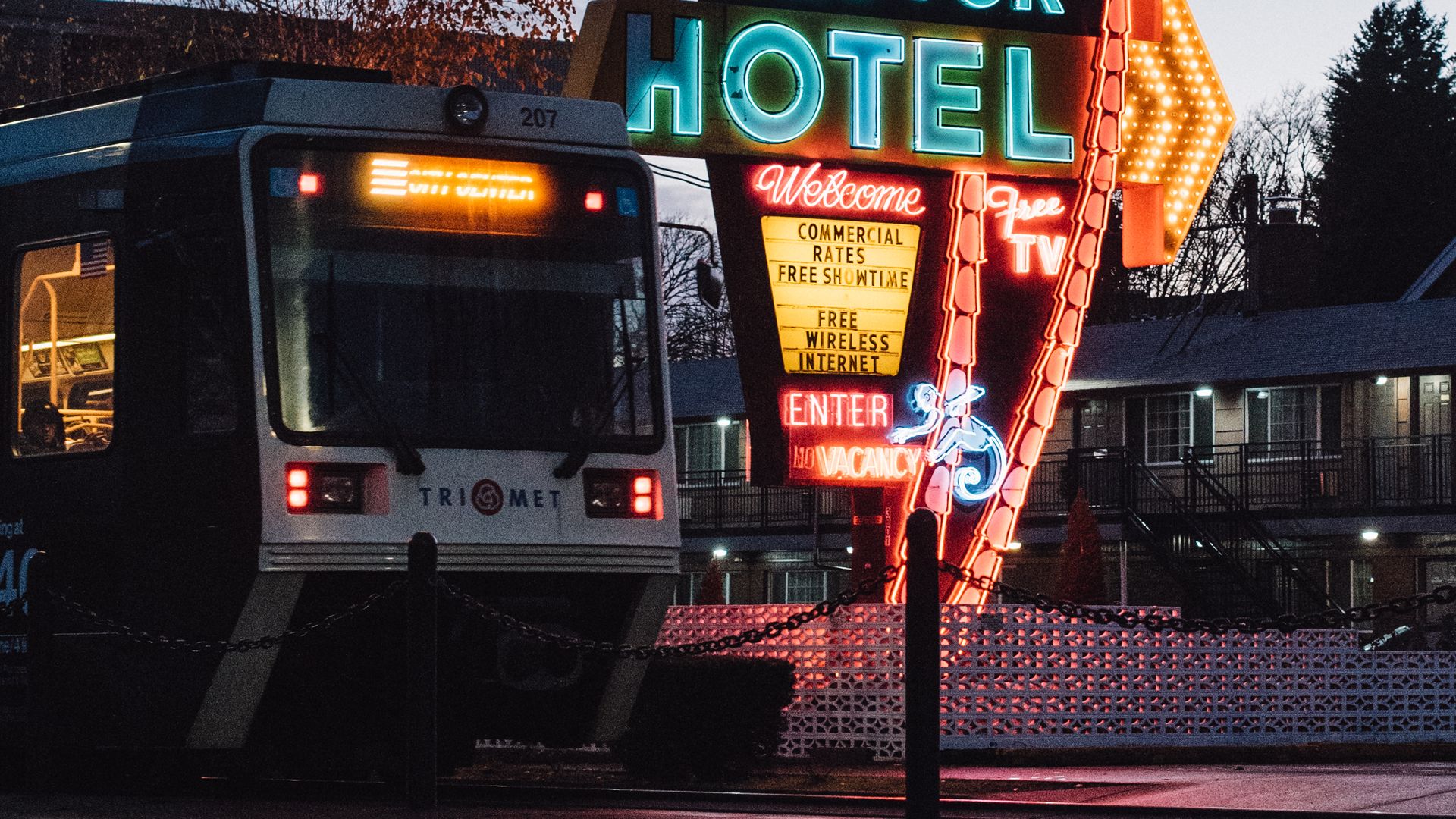 Night scene featuring a large neon palm-tree sign with "The Palms" in red script and turquoise "MOTOR HOTEL"; a bus in the foreground, power lines overhead, and bare trees against a dusky blue sky.