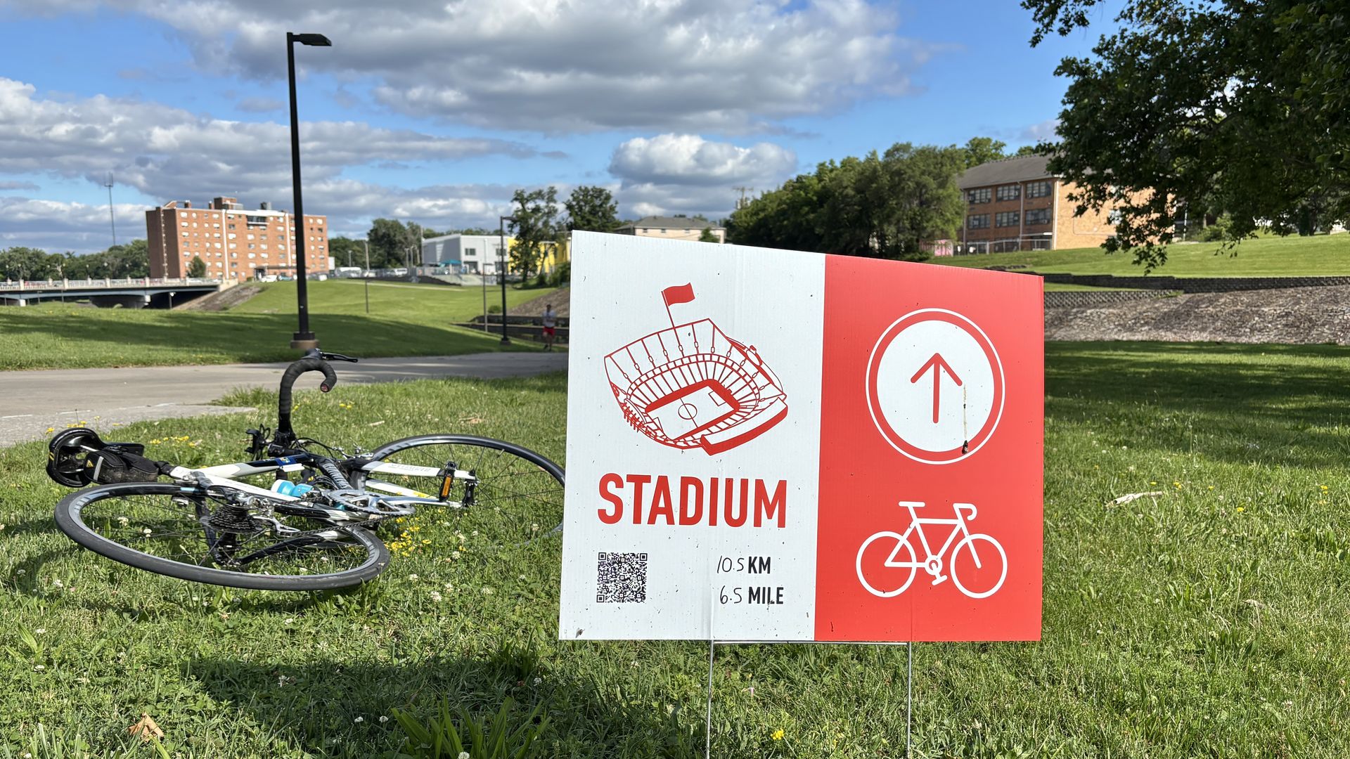Outdoor park scene with a red and white stadium sign in the foreground right, bicycles on the grass, a lamp post, a path, trees, and brick buildings under a blue sky with scattered clouds.