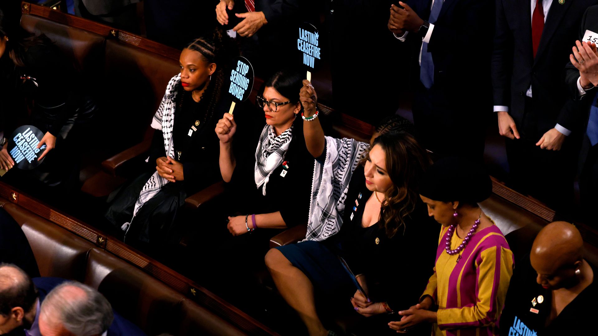 A group of female, POC members of Congress mostly wearing black and keffiyehs, holding up pro-Palestinian signs in the House chamber.
