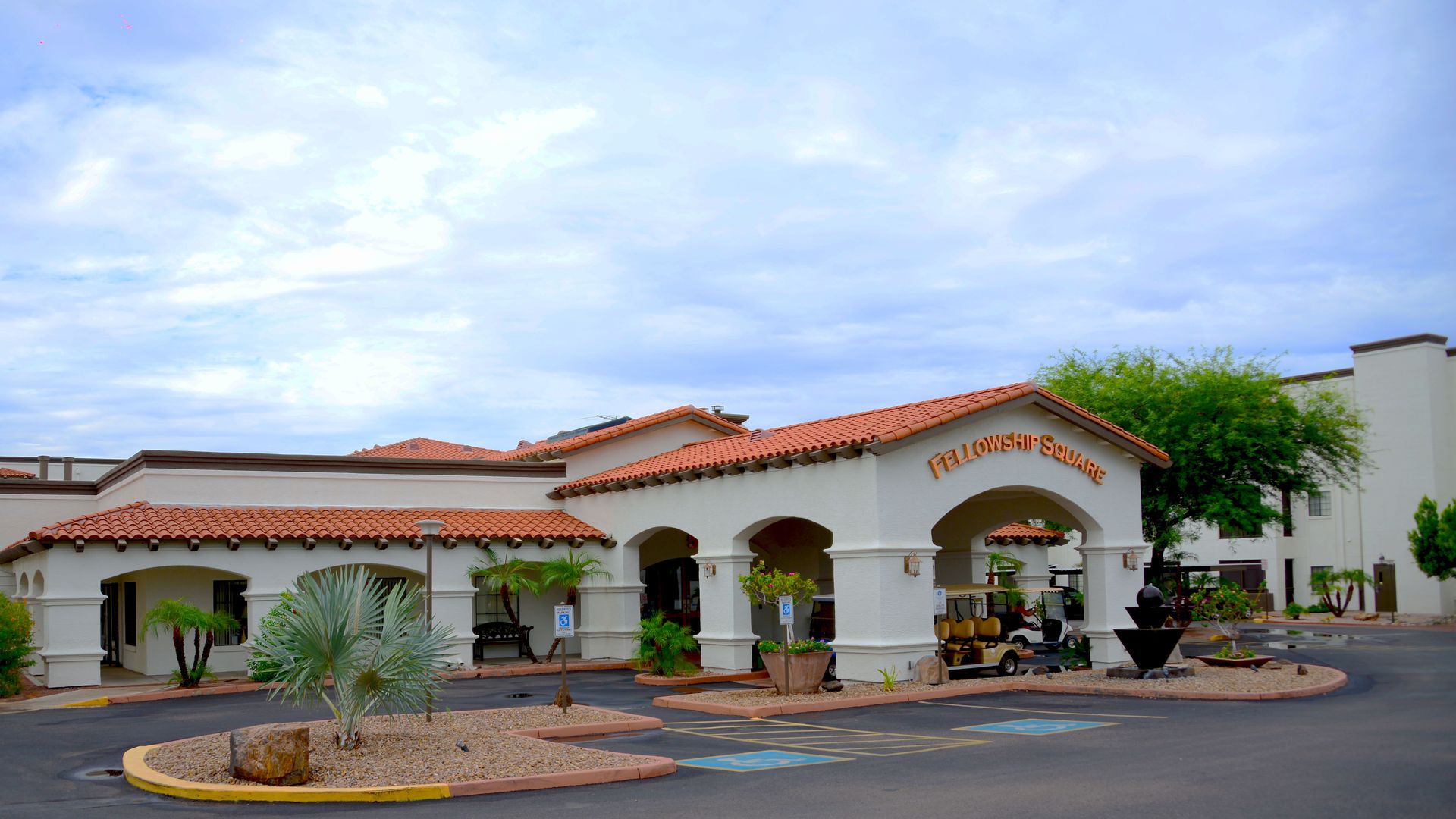 White stucco building with red tile roof and archway entrance labeled "Fellowship Square" under cloudy blue sky, surrounded by desert plants and handicap parking spaces.