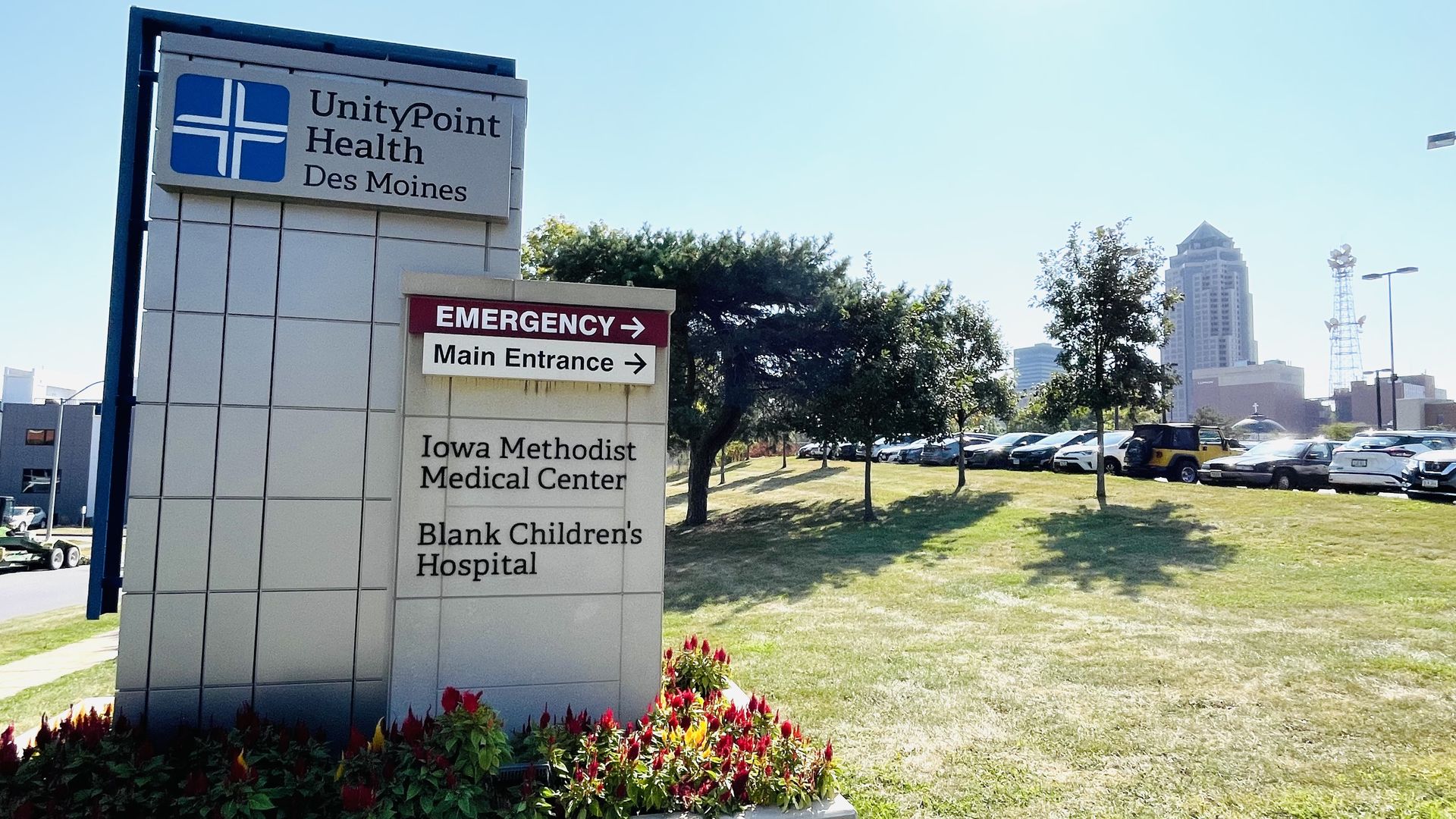 Sign for UnityPoint Health Des Moines with emergency and main entrance directions, located near a grassy area with flowers, trees, and a city skyline in the background under a clear sky.