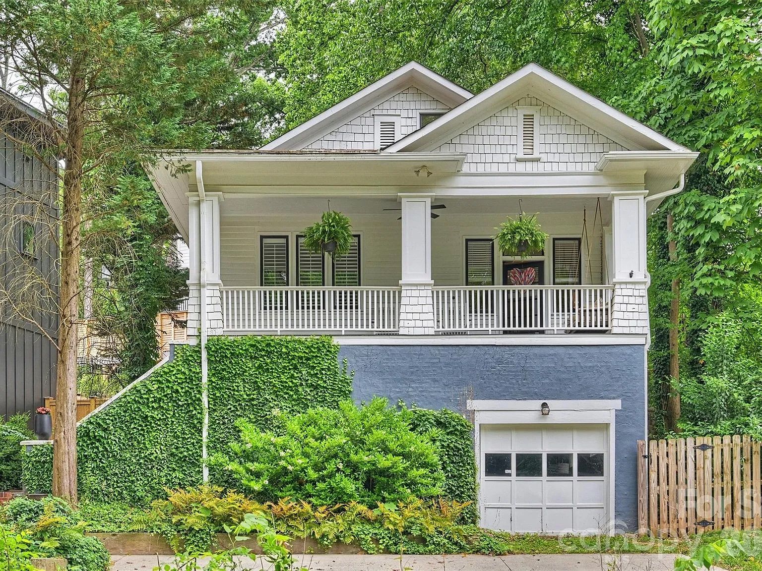 White craftsman-style house with a blue-gray raised foundation, a wide front porch, white railing and columns, two hanging ferns, ivy on the lower wall, and lush green trees surrounding.