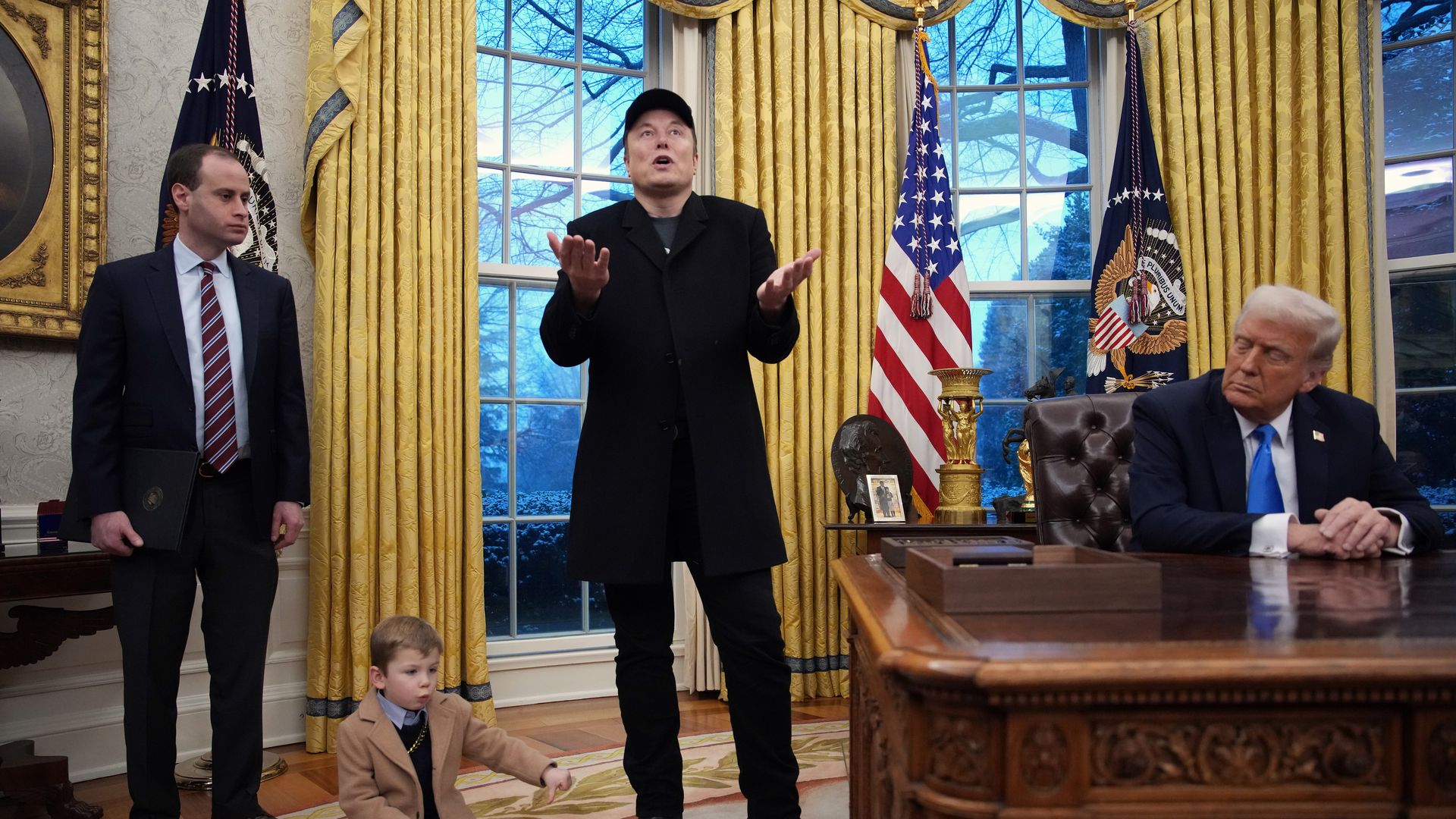 Elon Musk is at the center of the image, standing and speaking, with his son on the floor next to him and Donald Trump seated at a desk to his other side. 
