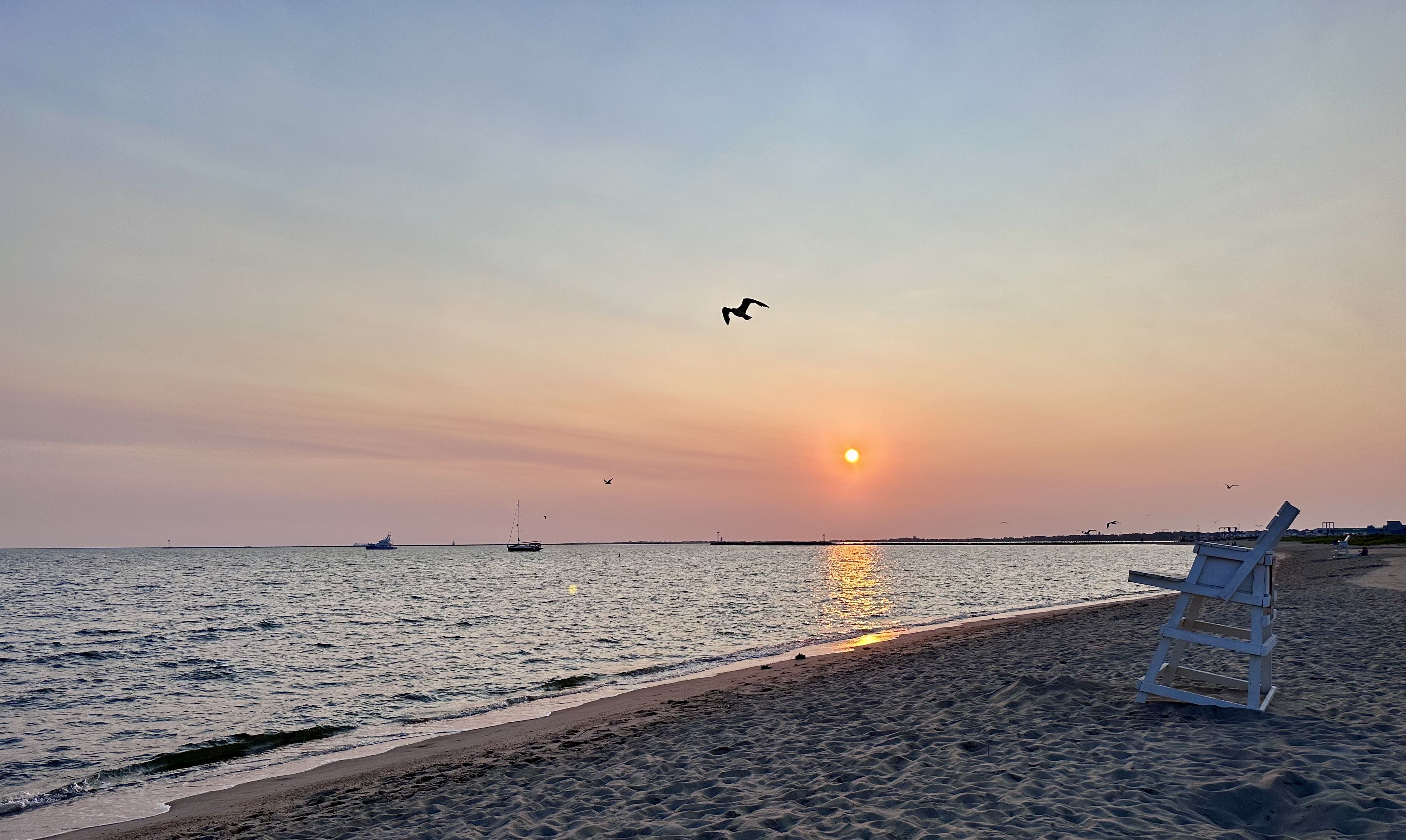 Beach at sunset with orange and blue sky, calm water, sailboats on horizon, a lifeguard chair on the sand, and birds flying above.