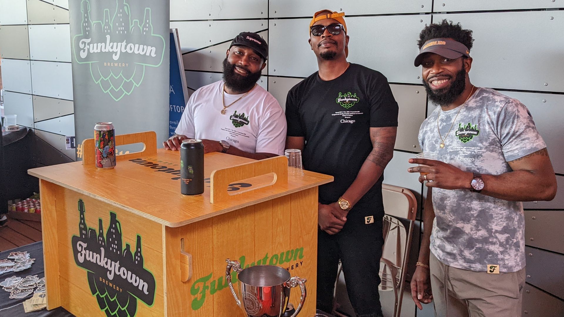 Wood box that reads "Funkytown" on front, with two beer cans on top. Three men standing behind box and smiling.