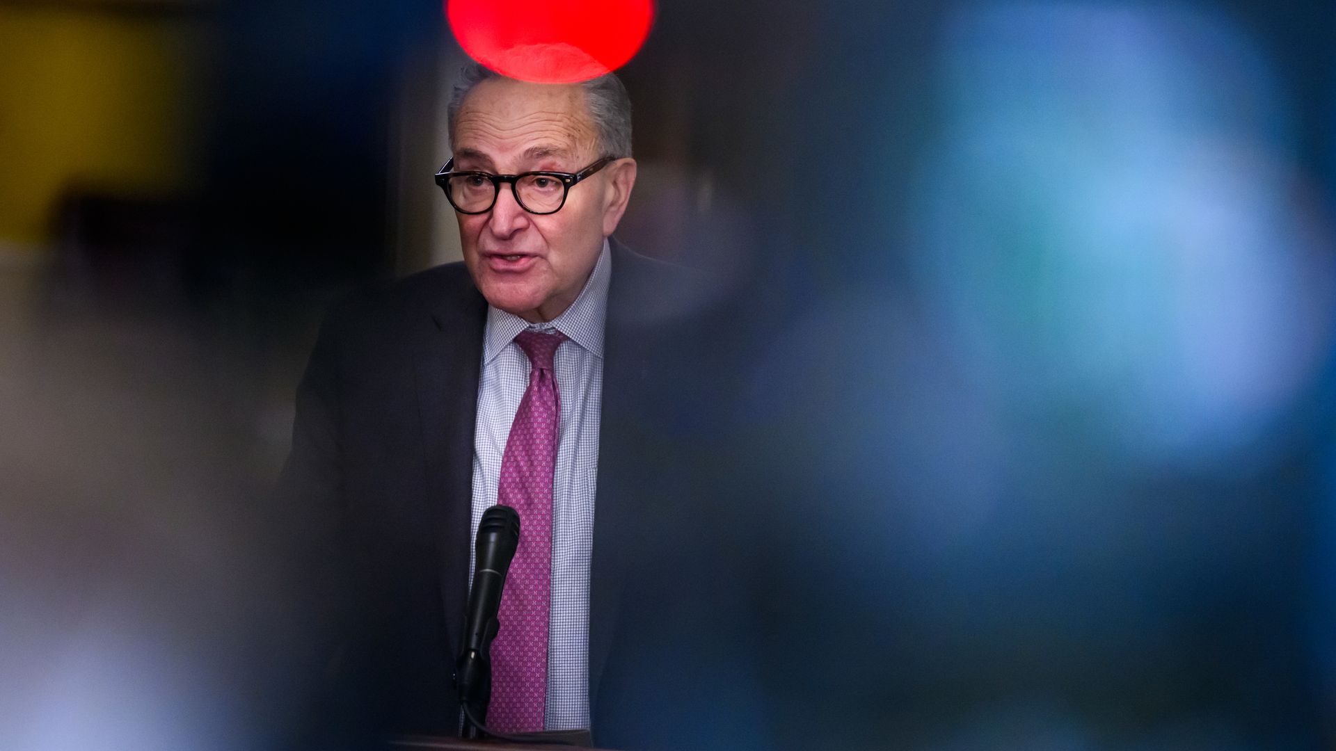 Sen. Chuck Schumer, wearing a gray suit, is seen in a yellow hallway with blurry camera equipment in the foreground.