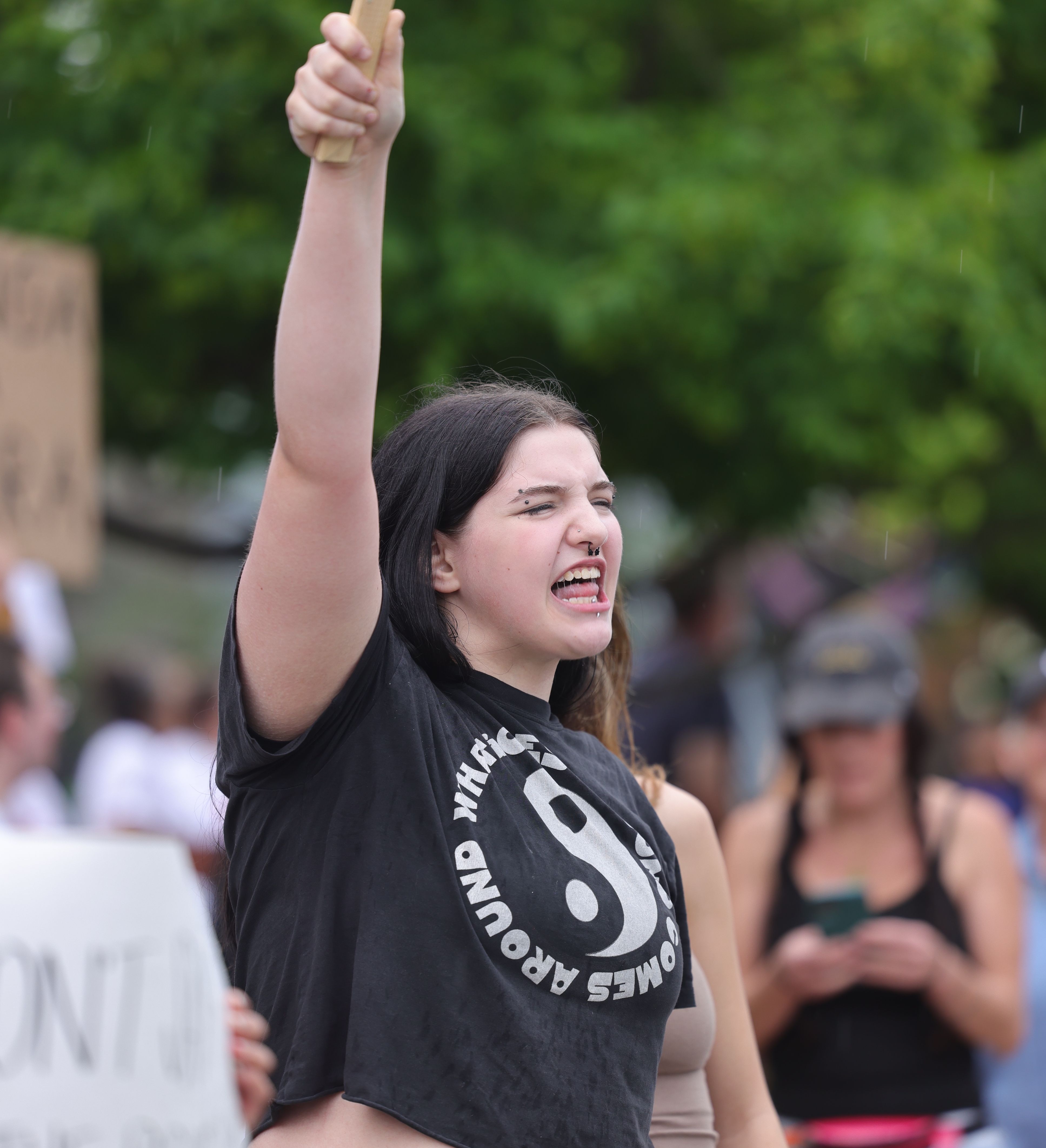 Young woman with piercings shouts passionately at a protest, holding a sign aloft in light rain, surrounded by demonstrators.