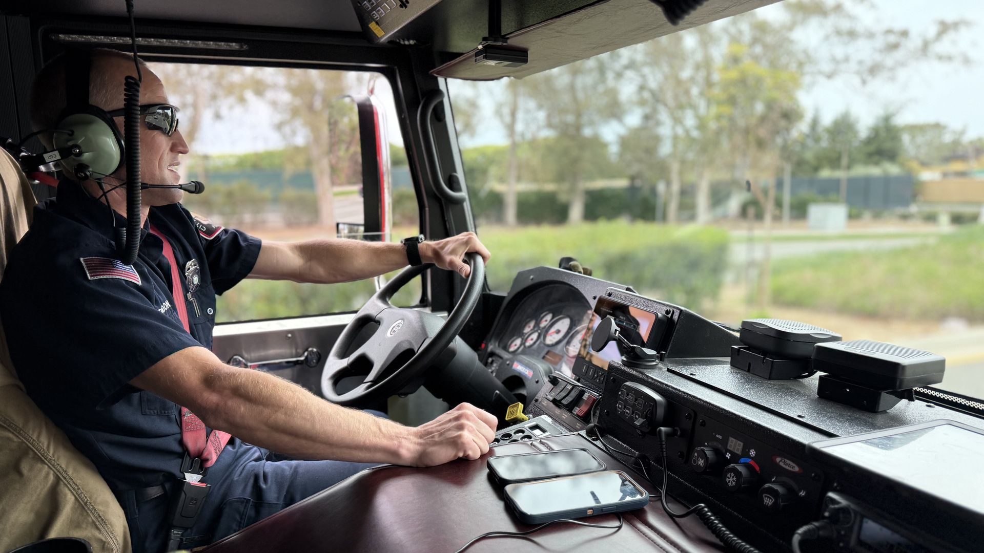 San Diego Fire-Rescue engineer Kevin Taddonio driver with headset and sunglasses sits at the wheel inside a cab, dashboard gauges and devices in view, blurred trees outside through the windshield.