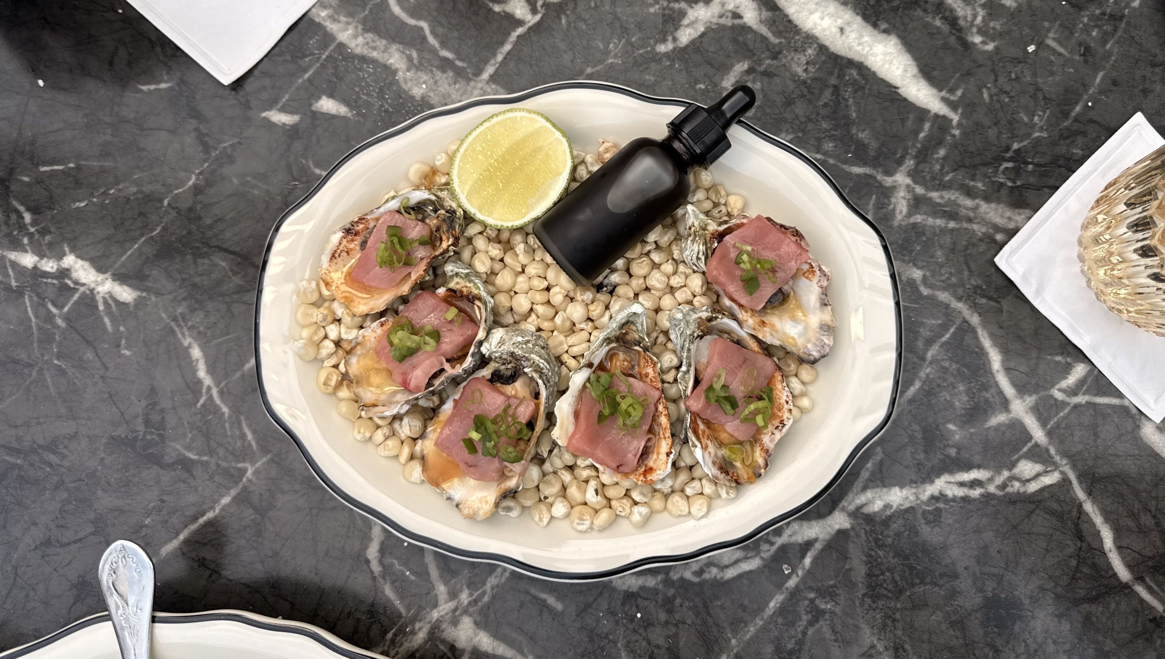 Top-down view of a white scalloped platter on dark marble: oysters on shells over white beans, topped with pink meat and green garnish. A lemon wedge and a dark dropper bottle sit center.