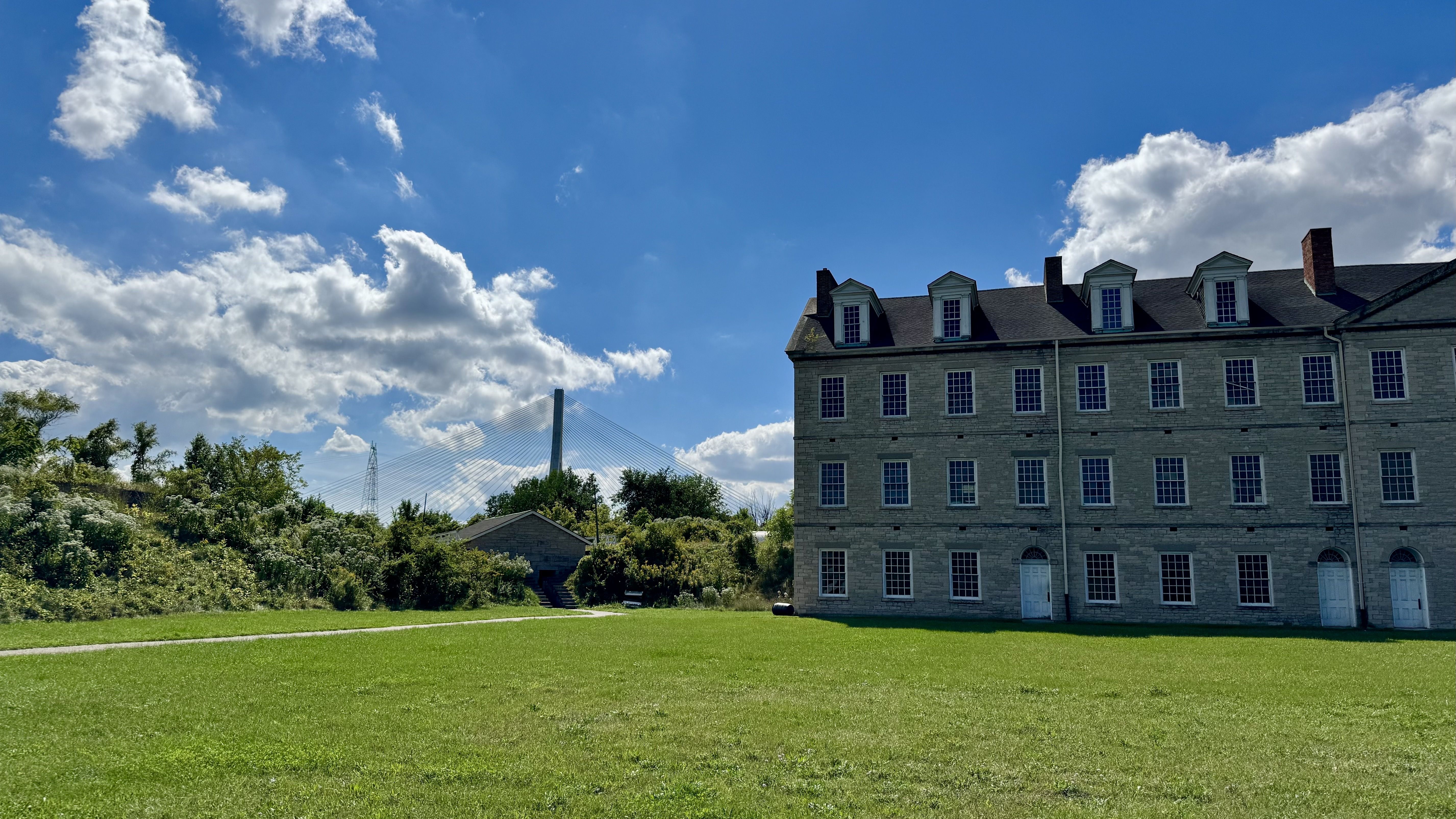A grassy field showing the edges of a fort, covered in greenery. Bridge visible far in the background. The building in the foreground is a limestone barracks.