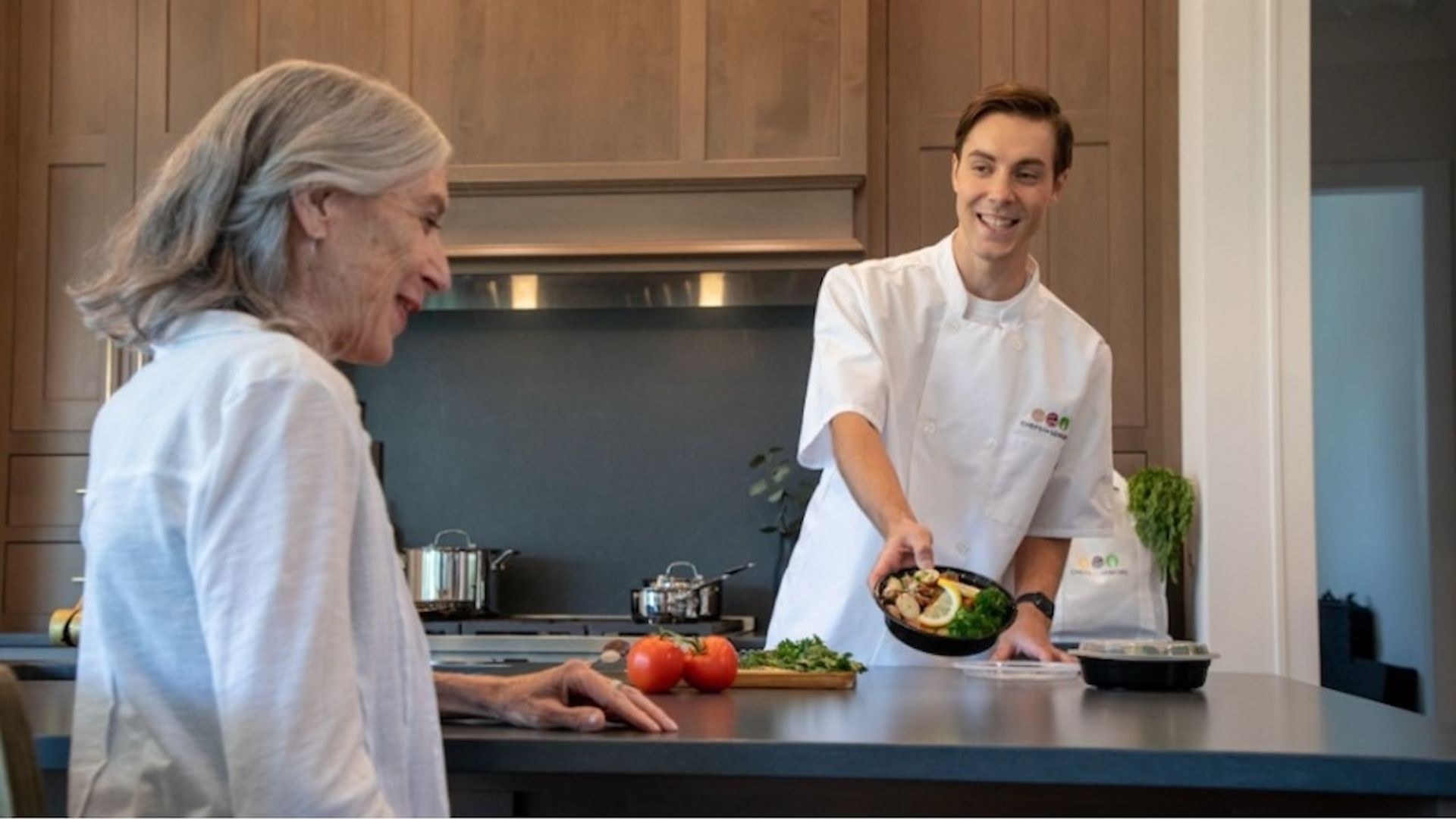 A chef and a woman are in a kitchen. The chef is holding a plate of food. 