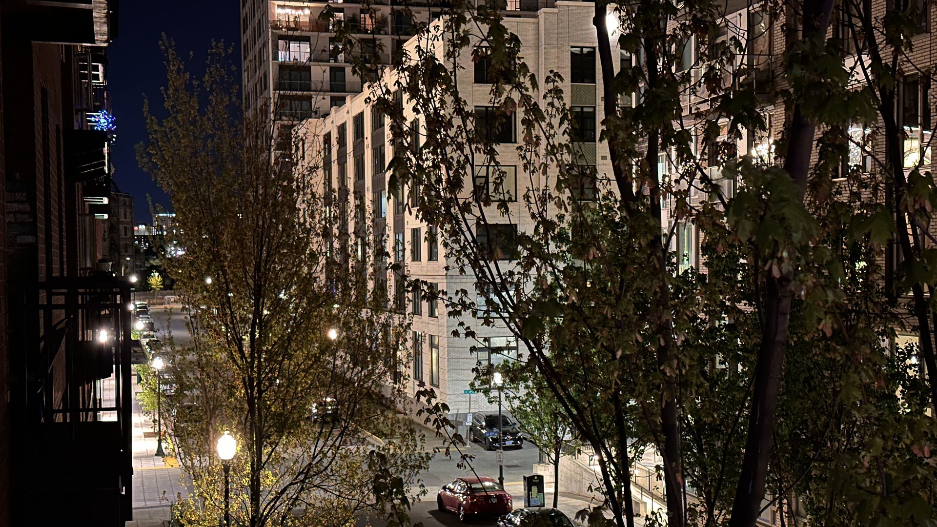 A street and tall residential buildings lit up at night through trees