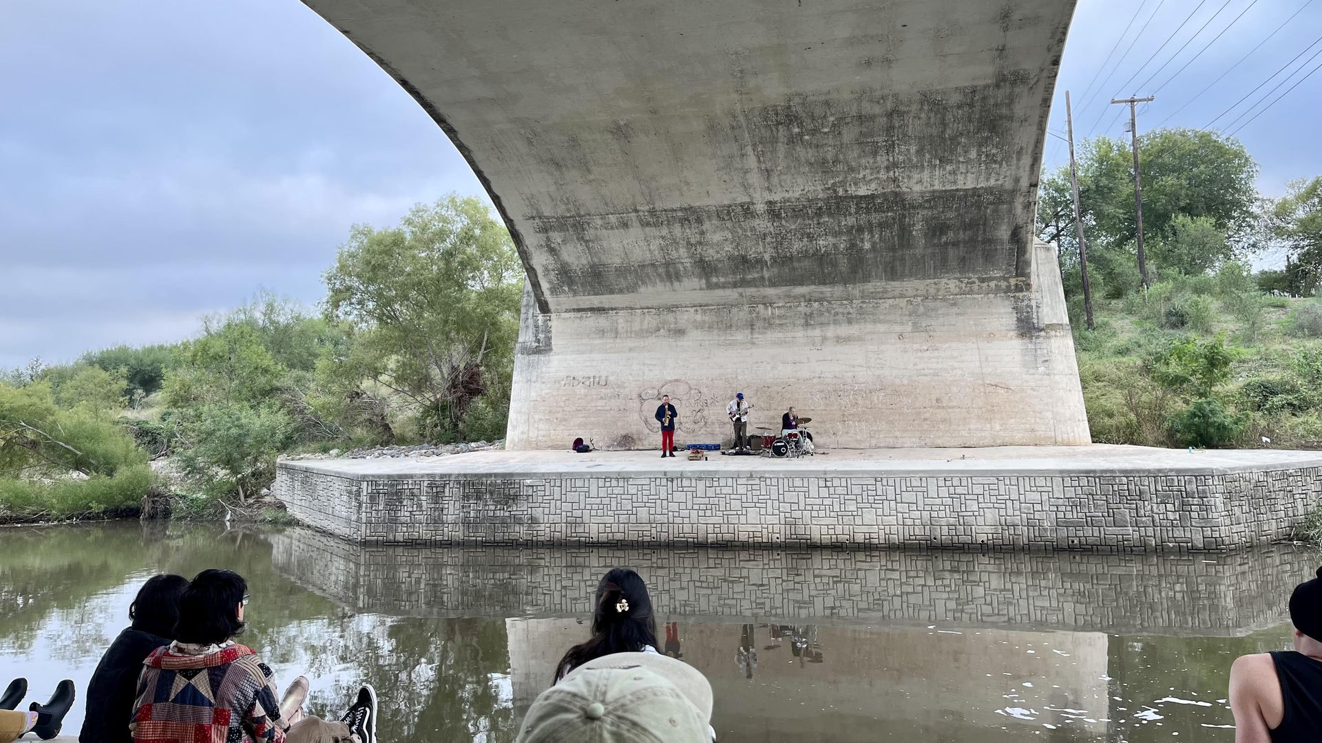 Three musicians performing under a large concrete bridge, called the Echo Bridge, near a calm river, with audience members sitting on the riverbank watching from across the San Antonio River, all surrounded by green trees and a cloudy sky.