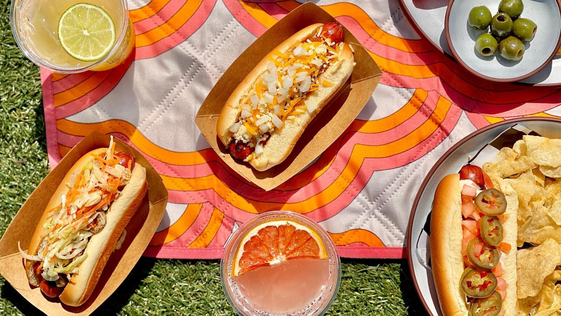 Picnic on a bright pink-orange blanket with two hot dogs topped with onions and cheese, olives, carrot sticks, chips, lemonade with lime, a grapefruit drink, and a dip bowl on the grass.