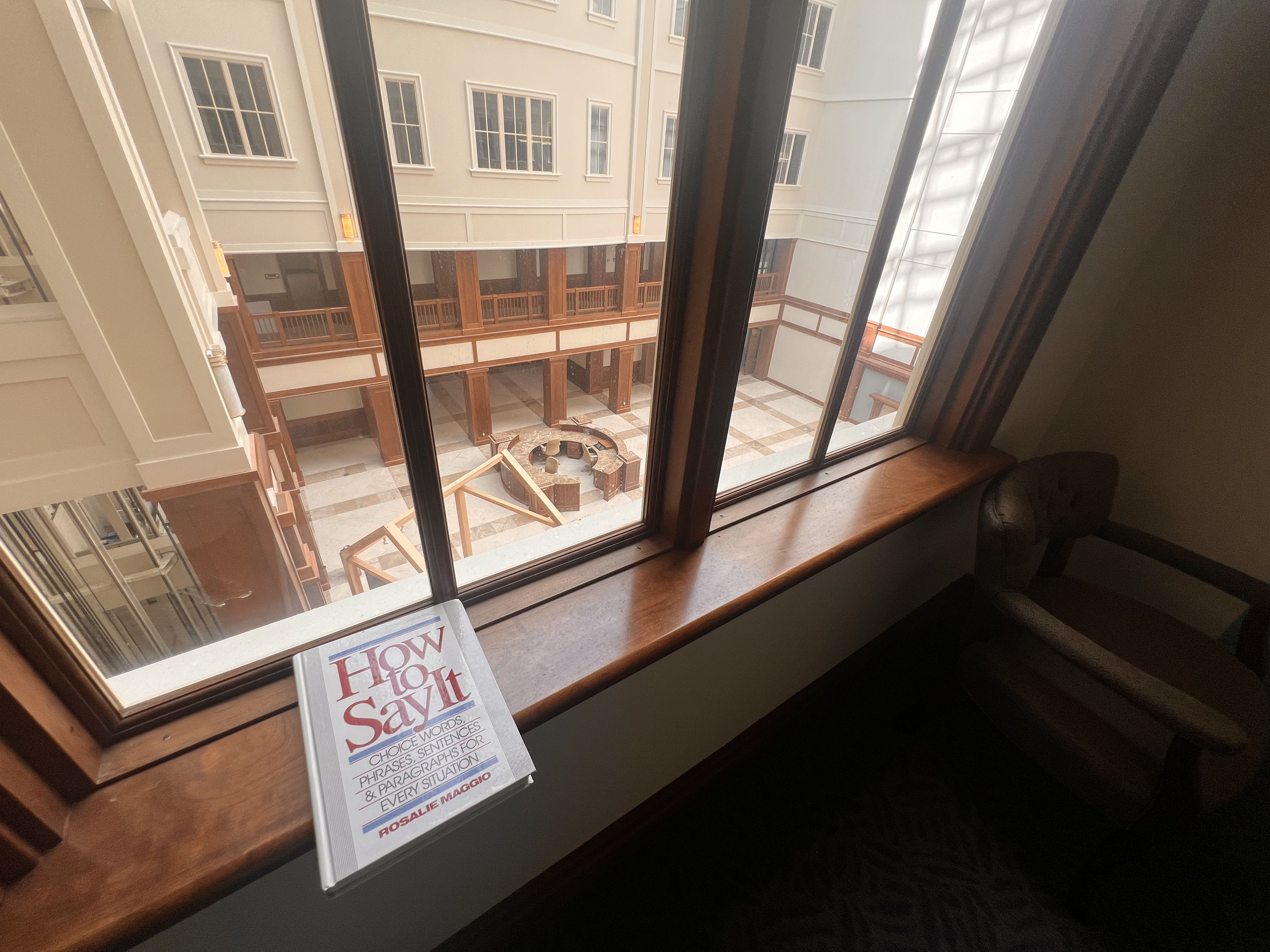 View from a window into an indoor atrium with beige walls, tall windows, and wooden railings around a circular fountain on a tiled floor; a magazine titled "How to Say It" rests on the sill.