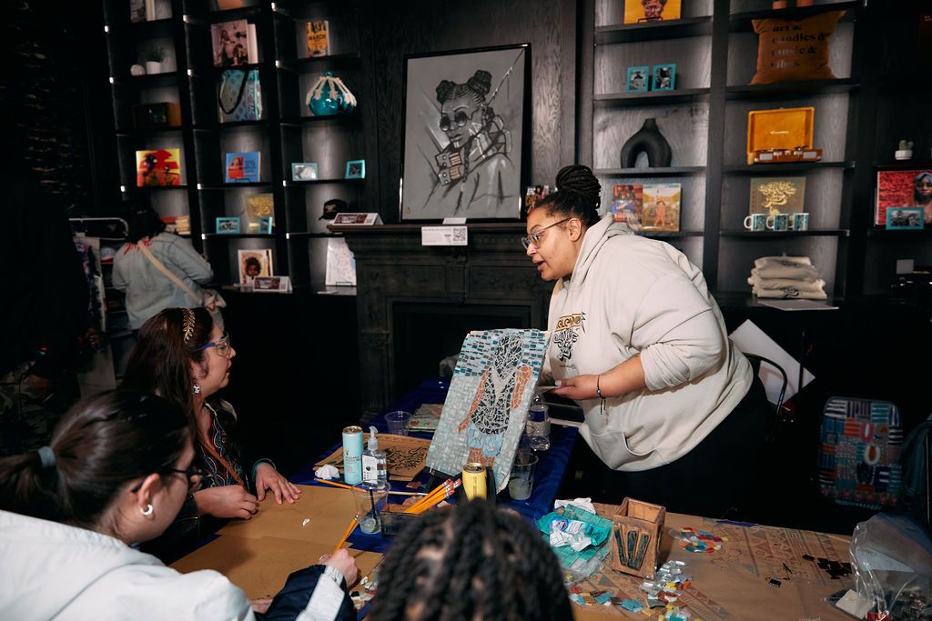 A woman in a gray hoodie holding an artwork and showing it to a person who sits at a table.