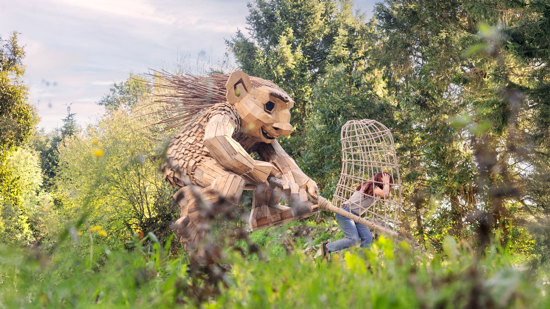 A large wooden horse sculpture made of interlocking planks sits in a sunny garden. A person in jeans stands nearby, with green trees and a lattice cage in the background.