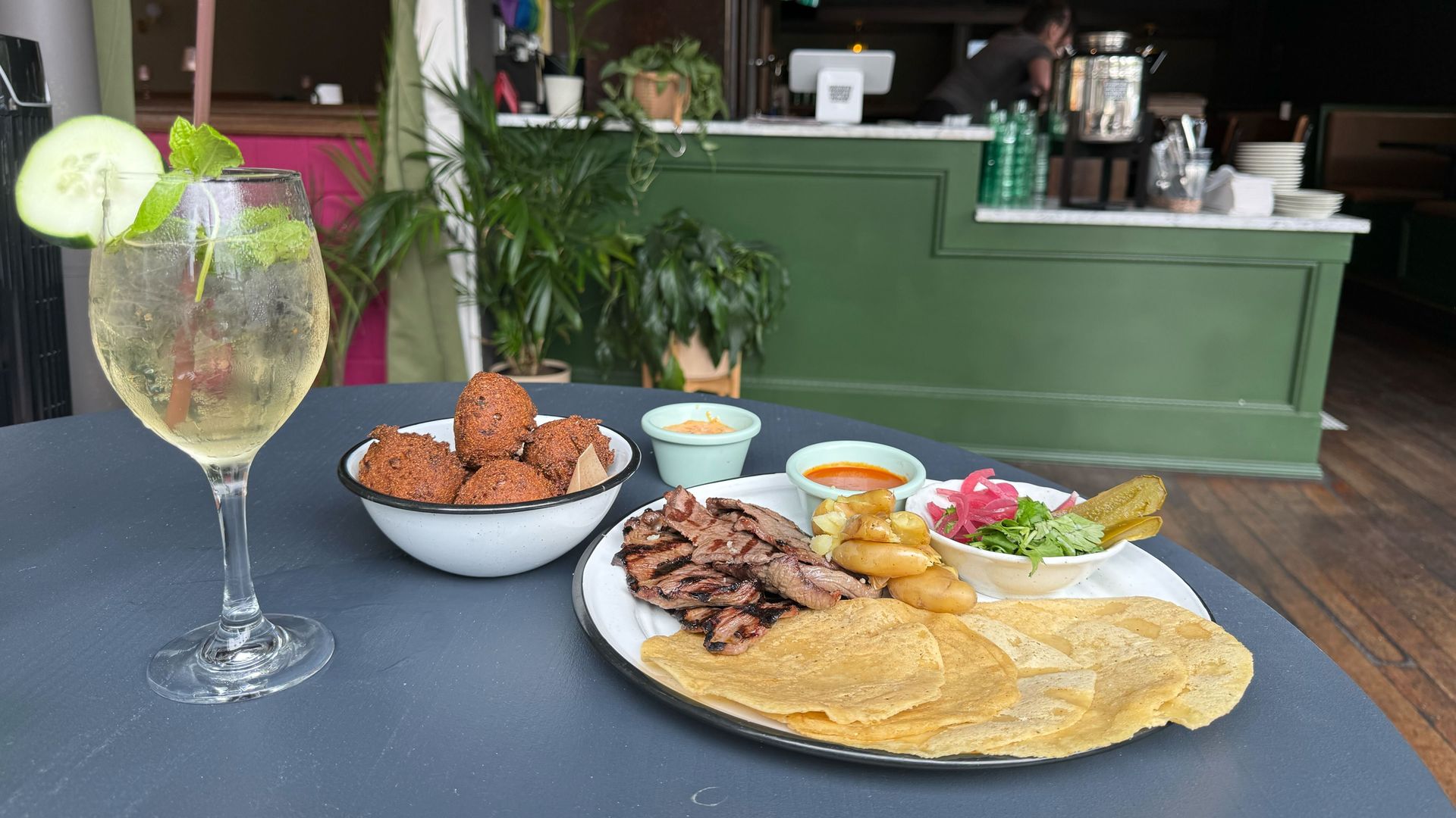 Table with a meal of grilled meat, potatoes, tortillas, pickled vegetables, sauces, and a bowl of fried balls, alongside a glass of iced drink garnished with cucumber and mint, in a cafe setting.