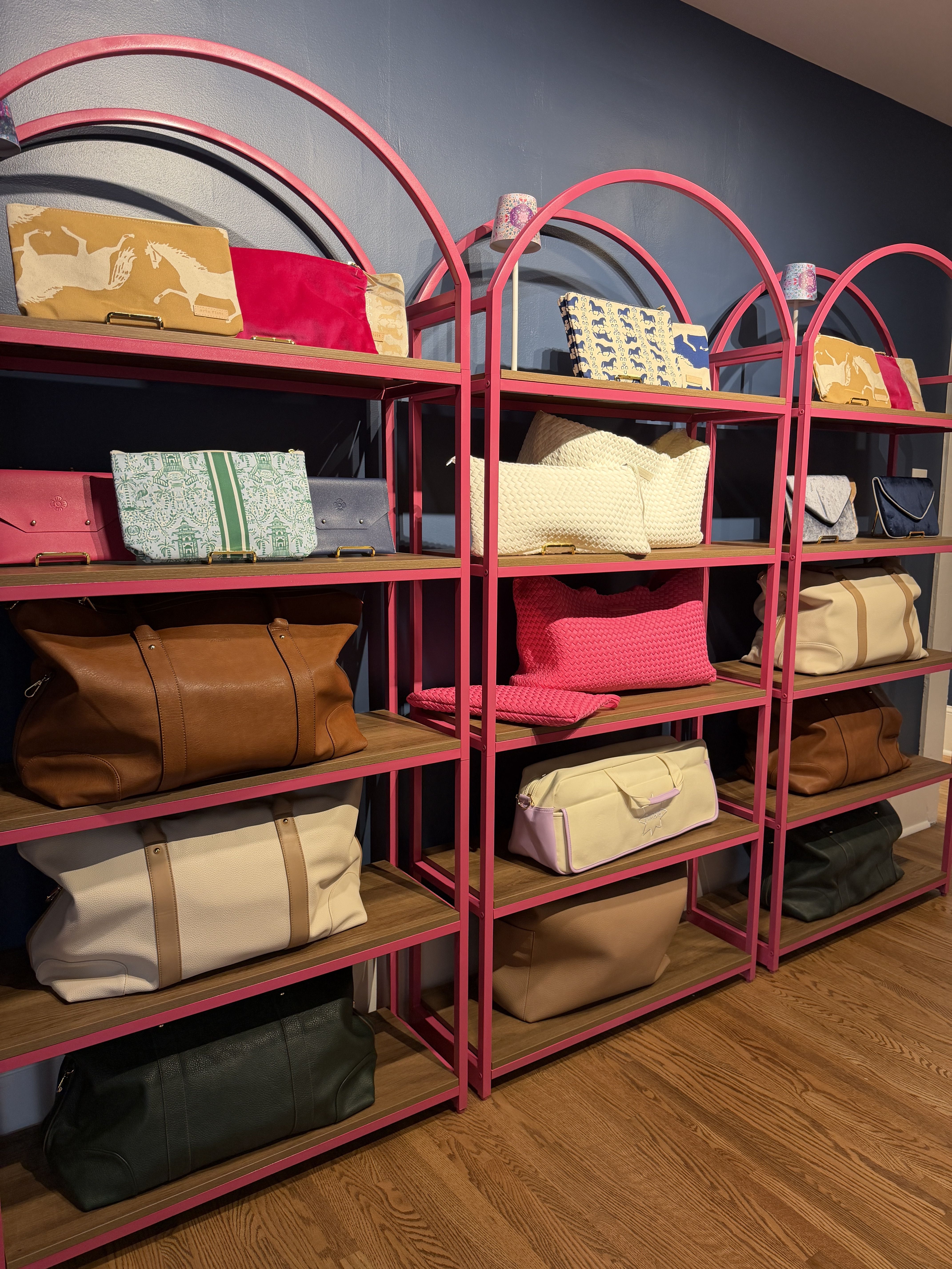 Boutique display featuring bright pink arched metal shelves filled with assorted handbags, totes, and clutches in various colors and patterns, set against a blue wall.