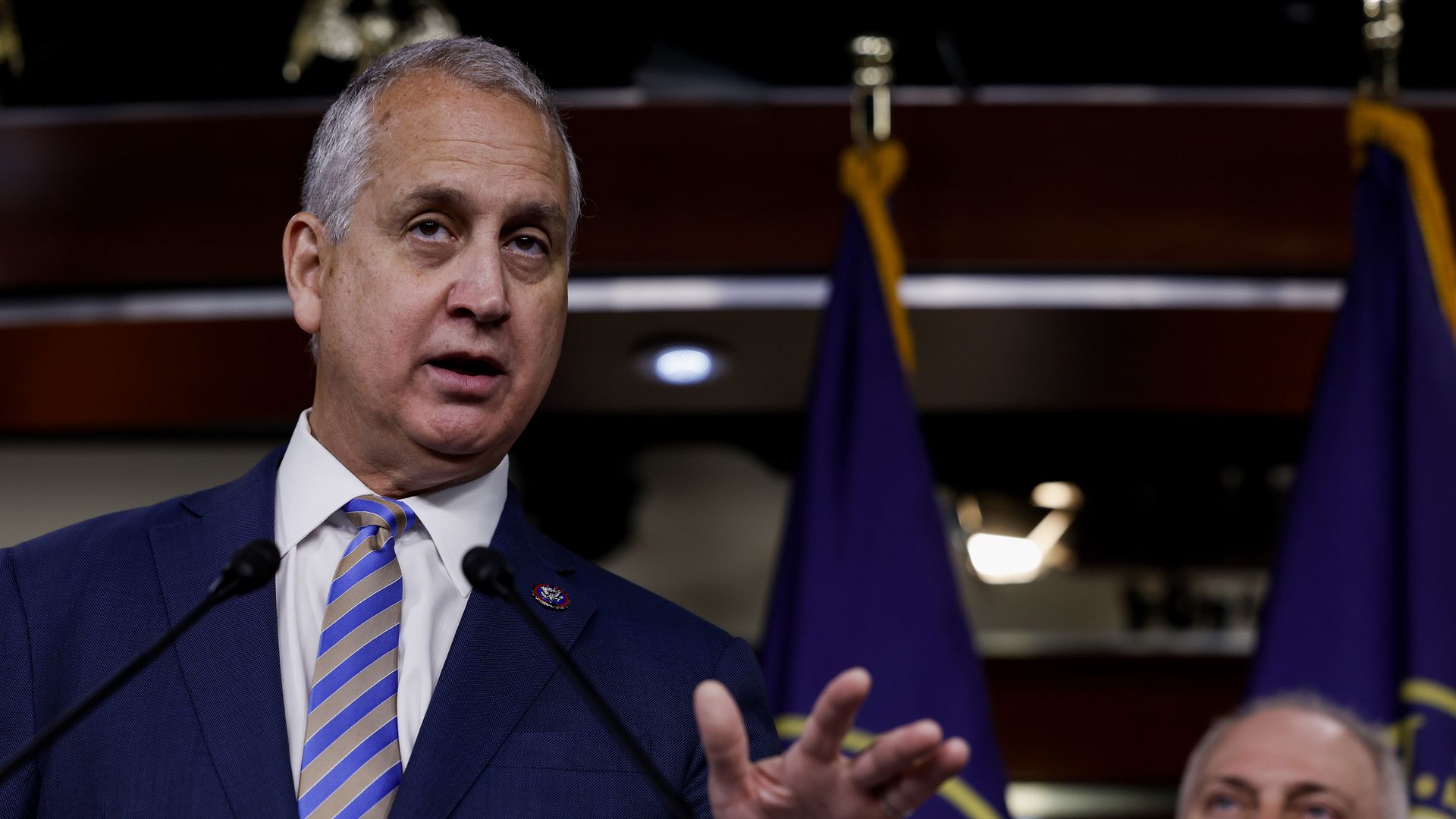 Rep. Mario Diaz Balart, wearing a blue suit and speaking at a press conference in front of blue flags.