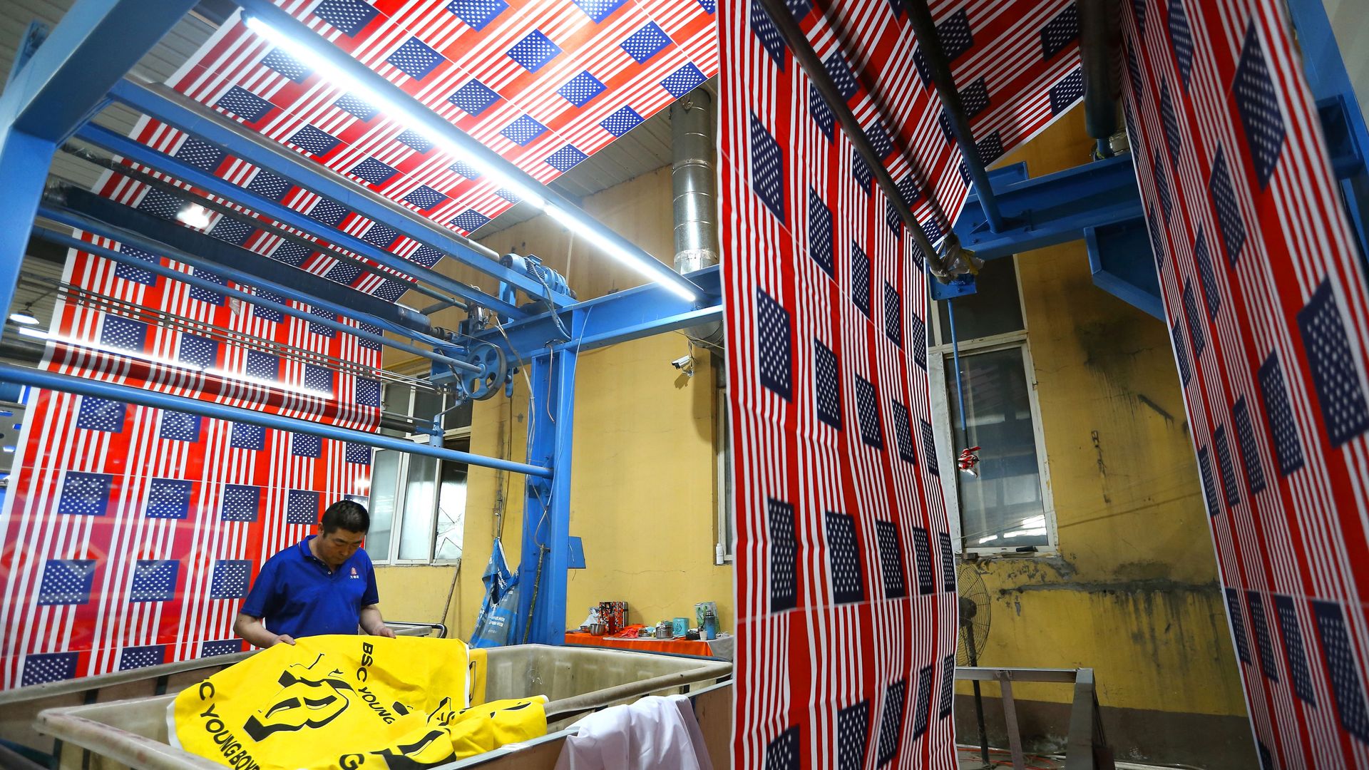 Industrial workshop with blue steel framework and hanging red, white, and blue American flag banners covering walls and ceiling; a worker in a blue shirt stands near yellow equipment.