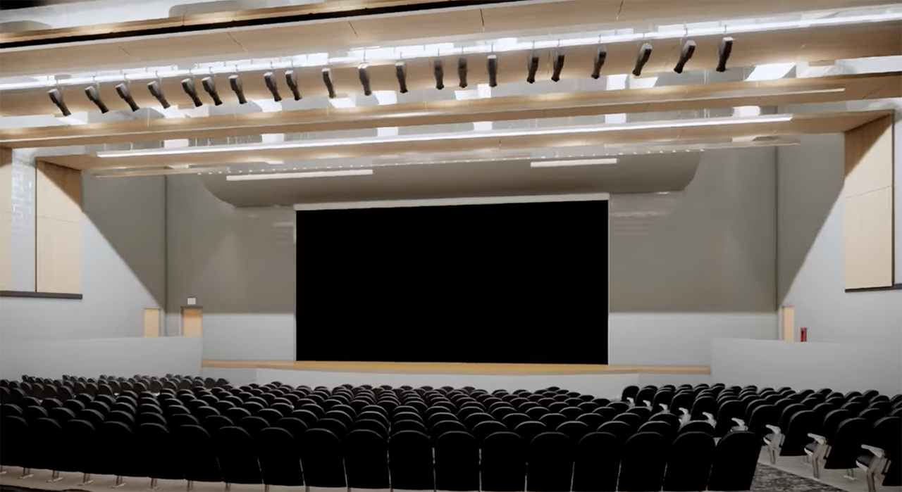 Empty auditorium with rows of black seats facing a large stage framed by light wooden panels and gray walls, under ceiling lights and structural beams.