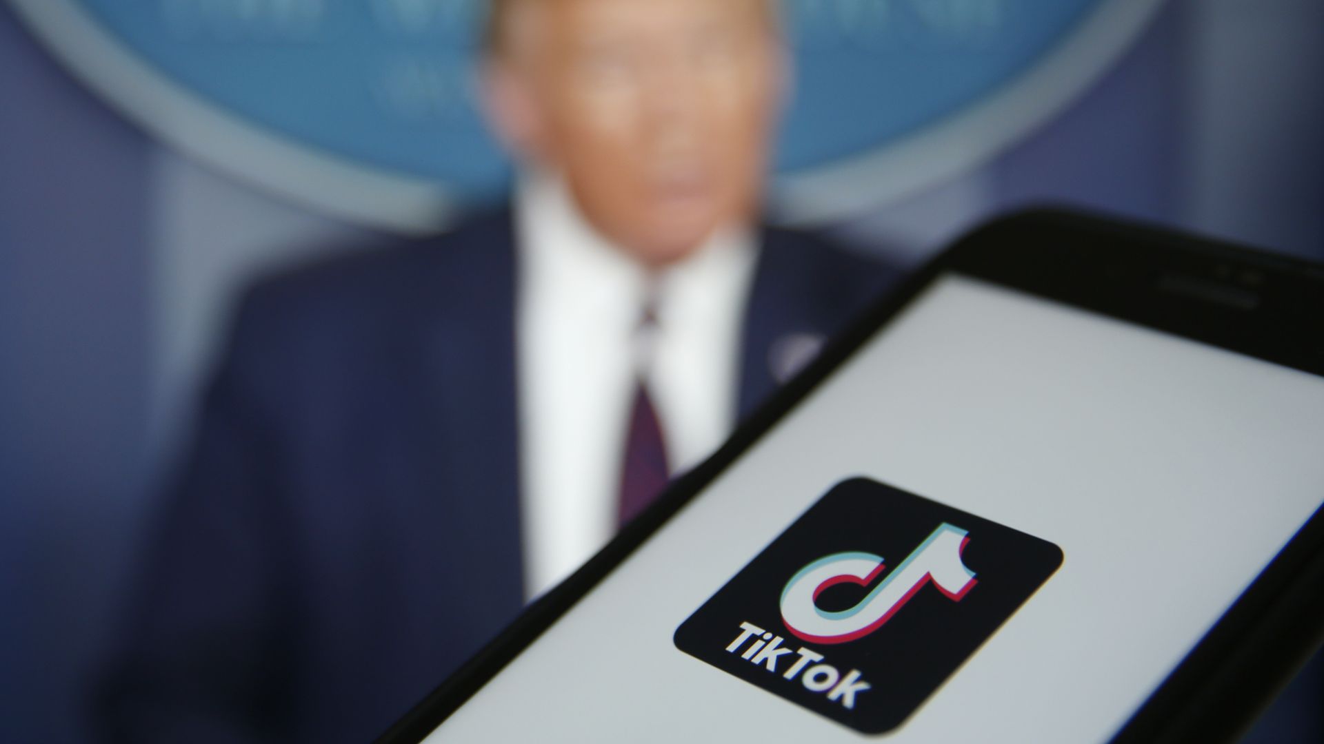 The TikTok logo is displayed in the app store in this arranged photograph in view of a video feed of U.S. President Donald Trump on Monday, Aug. 3, 2020.