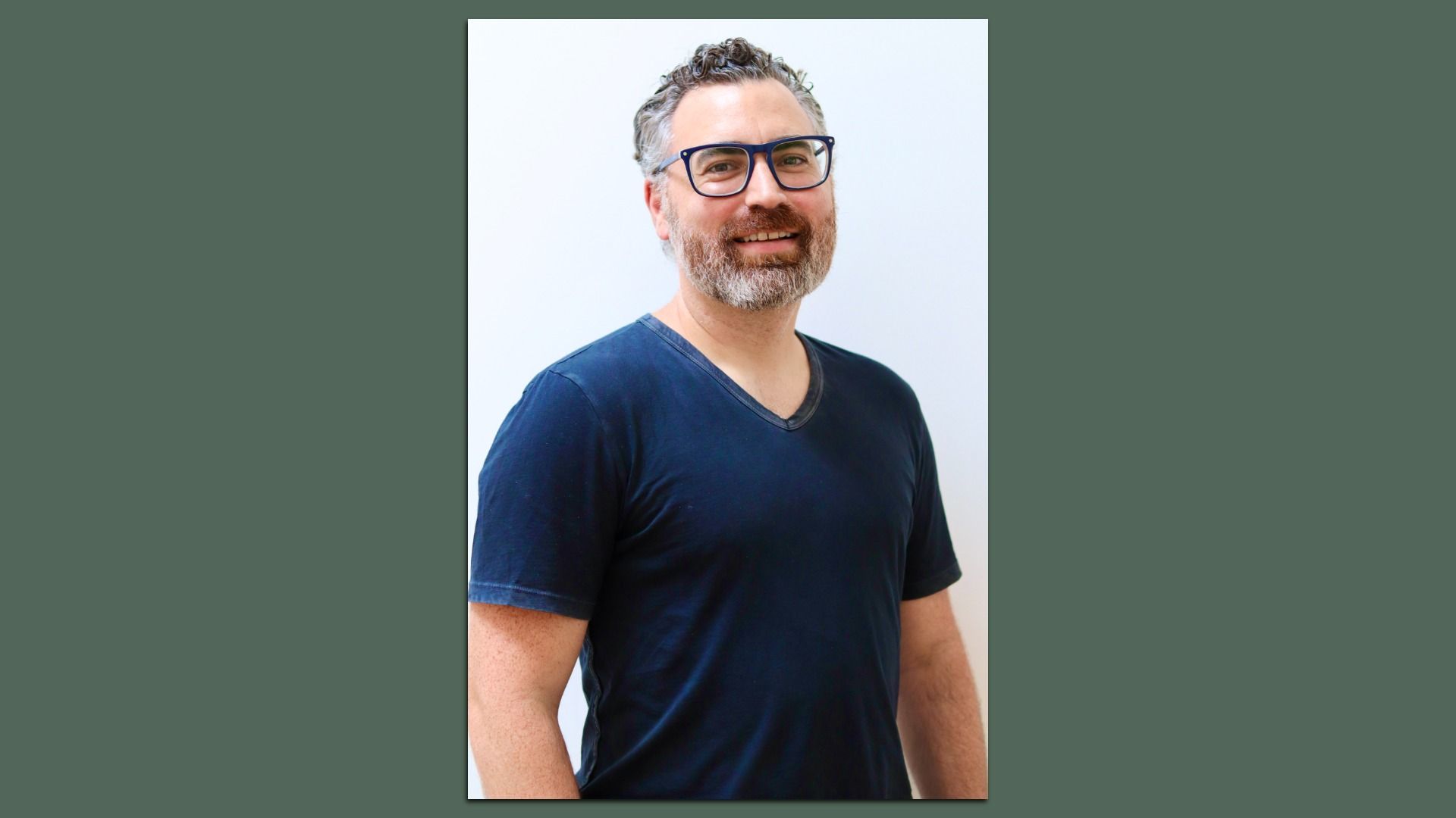 Smiling, middle-aged man with salt-and-pepper hair and beard, wearing a dark blue V-neck T-shirt and dark-framed glasses, standing against a pale background.