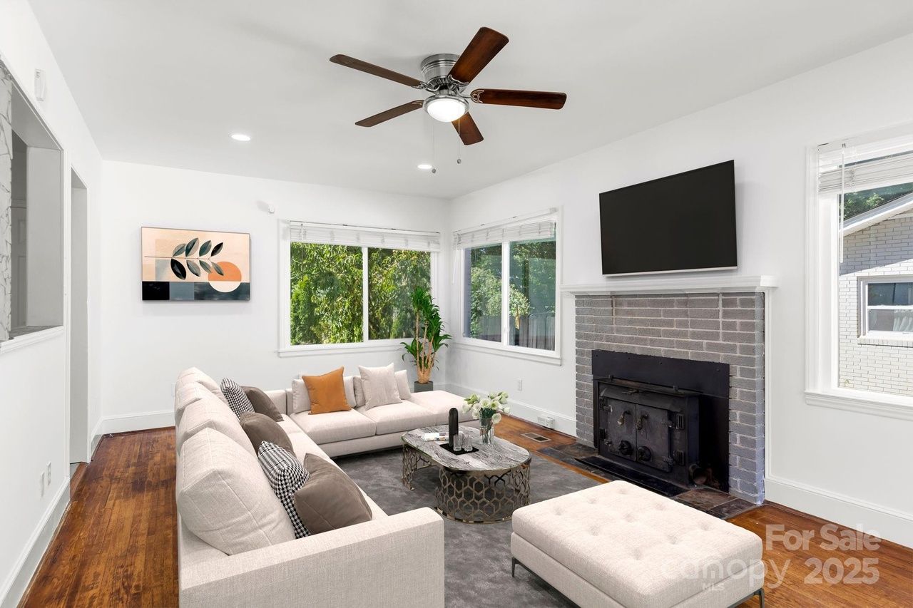 Bright living room with white walls, beige sectional sofa, patterned cushions, gray rug, wooden floor, ceiling fan, modern coffee table, large windows, and a gray brick fireplace with TV above.