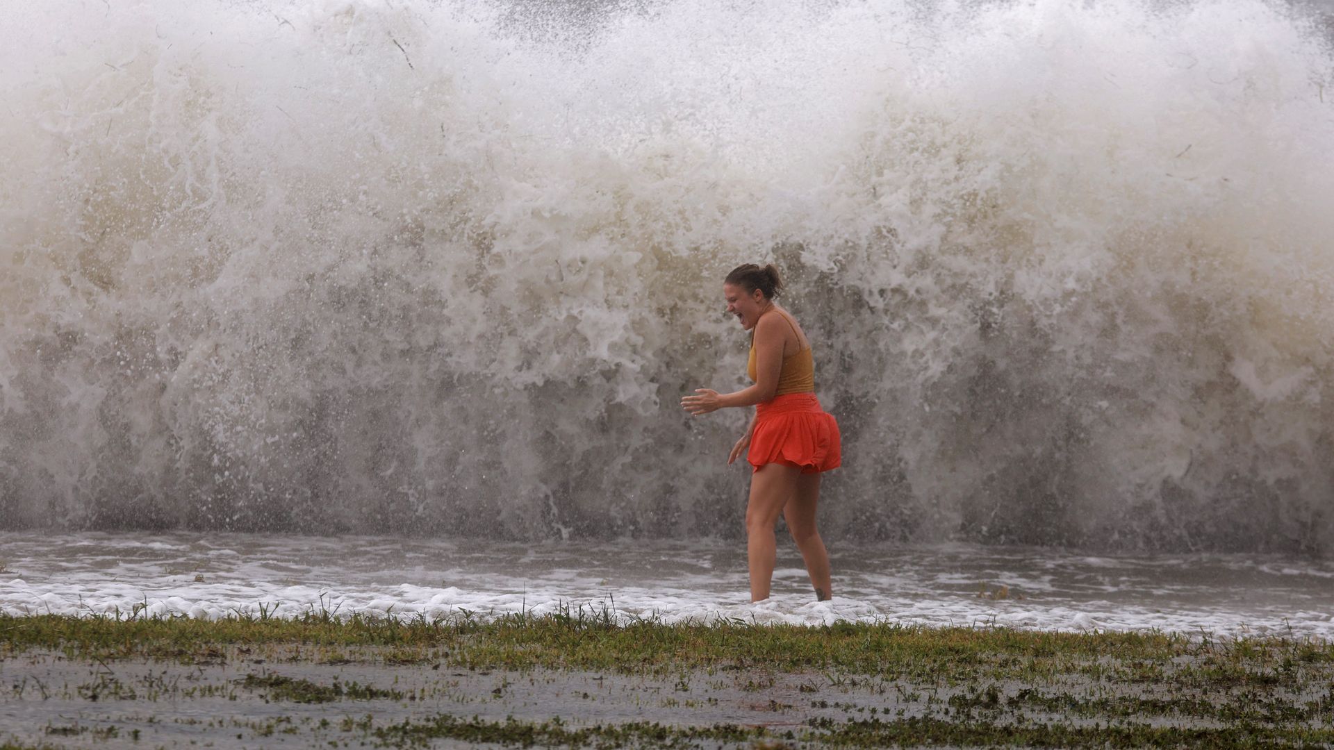 A wall of water behind a woman in shallow water in red shorts.