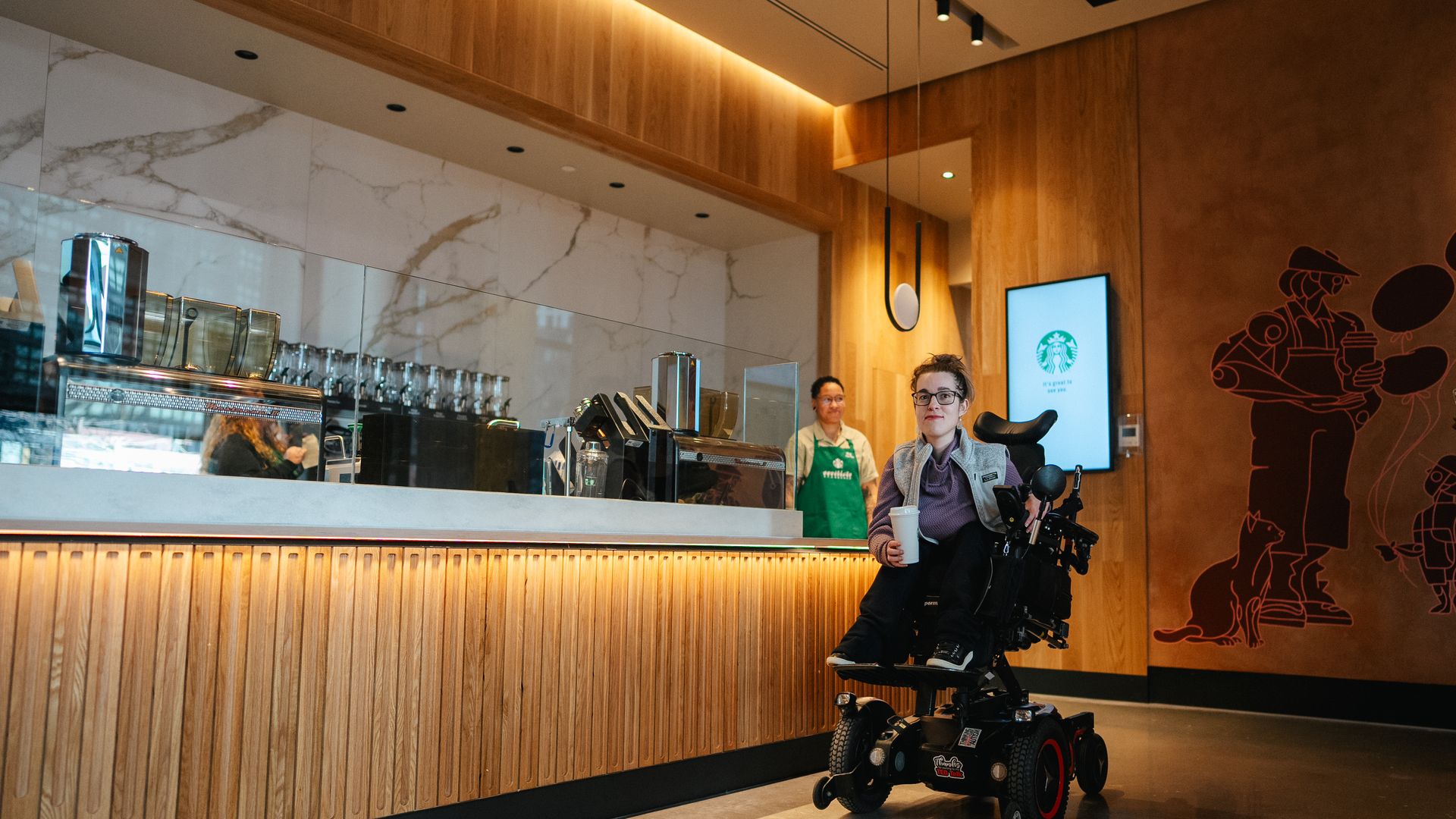 A woman in a wheelchair at the new Starbucks near Union Market with a backdrop of blonde wood features, an order status screen and employee in a green apron